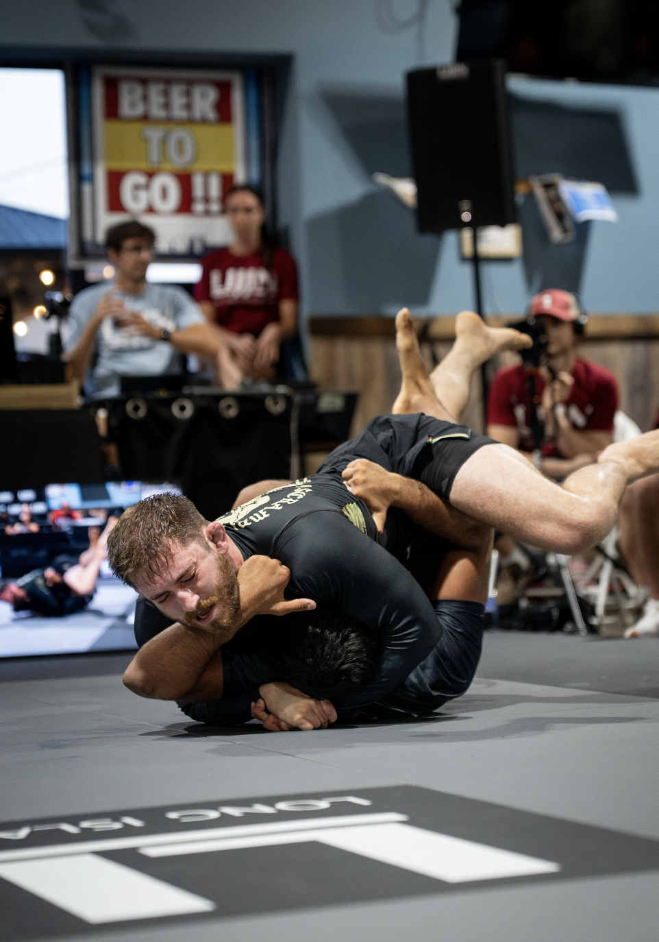 Two men are grappling on the mat during a Brazilian Jiu-Jitsu match, with one man in black dominating the other in a submission hold.