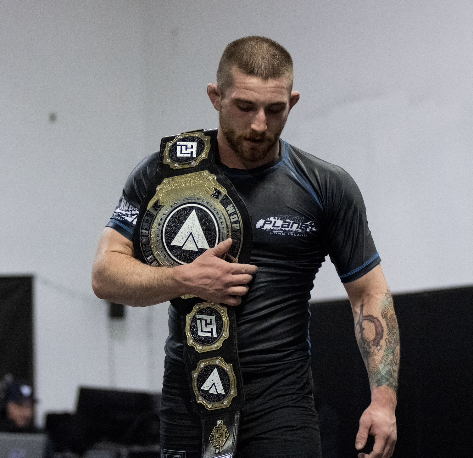 Man wearing a championship belt over his shoulder, looking down, dressed in black athletic wear, tattoos on his arm.