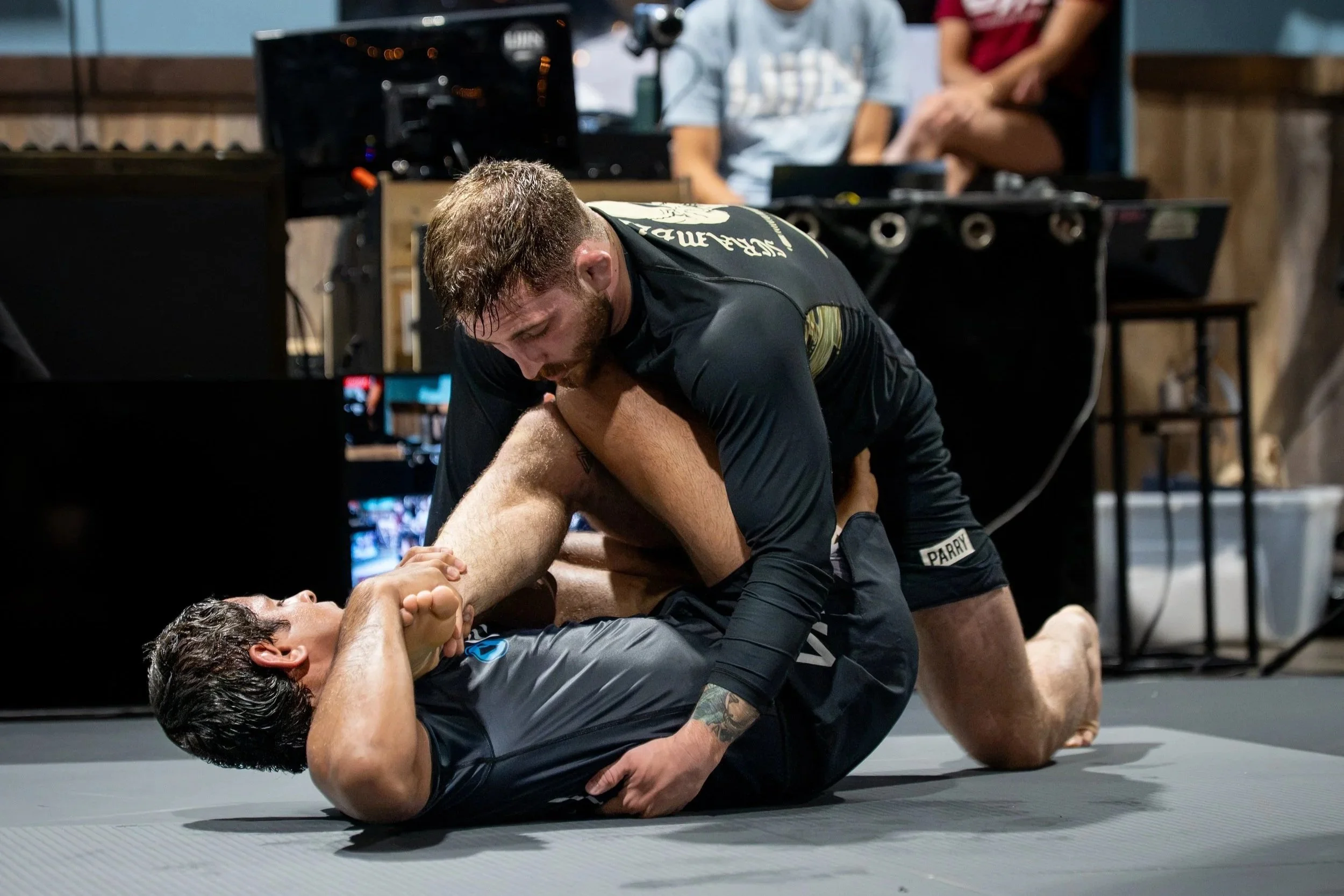 Two men are engaged in a Brazilian Jiu-Jitsu match on a mat, with one man on his back and the other on top, holding him and attempting a submission move. In the background, there are other people, equipment, and furniture in the gym or training facil