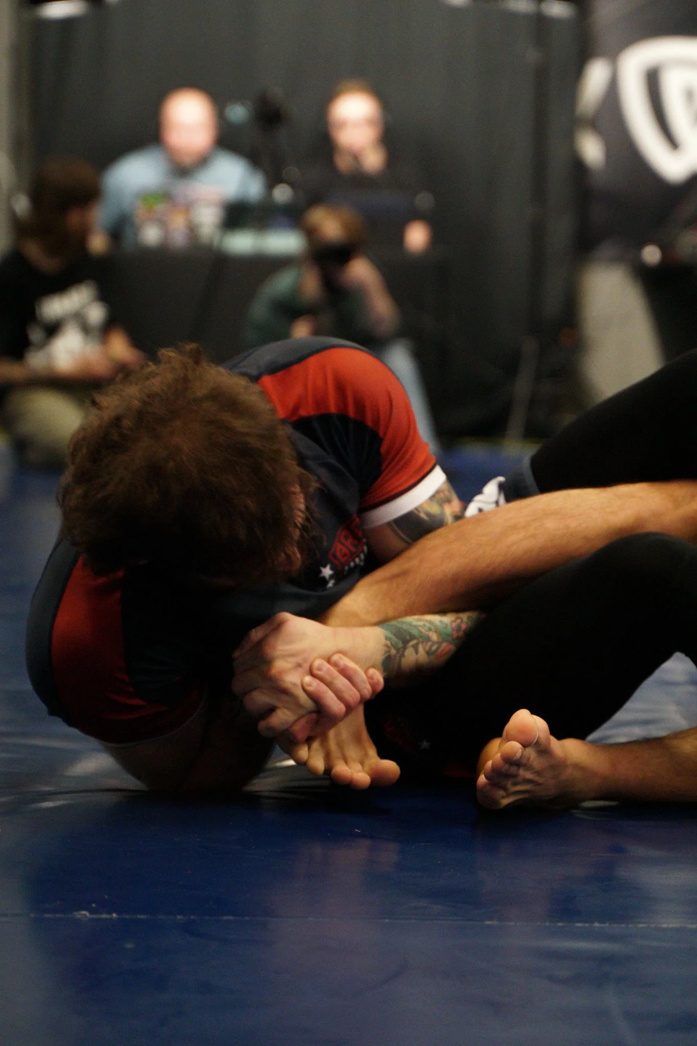 Dylan in a grappling hold on a blue mat, with spectators blurred in the background.