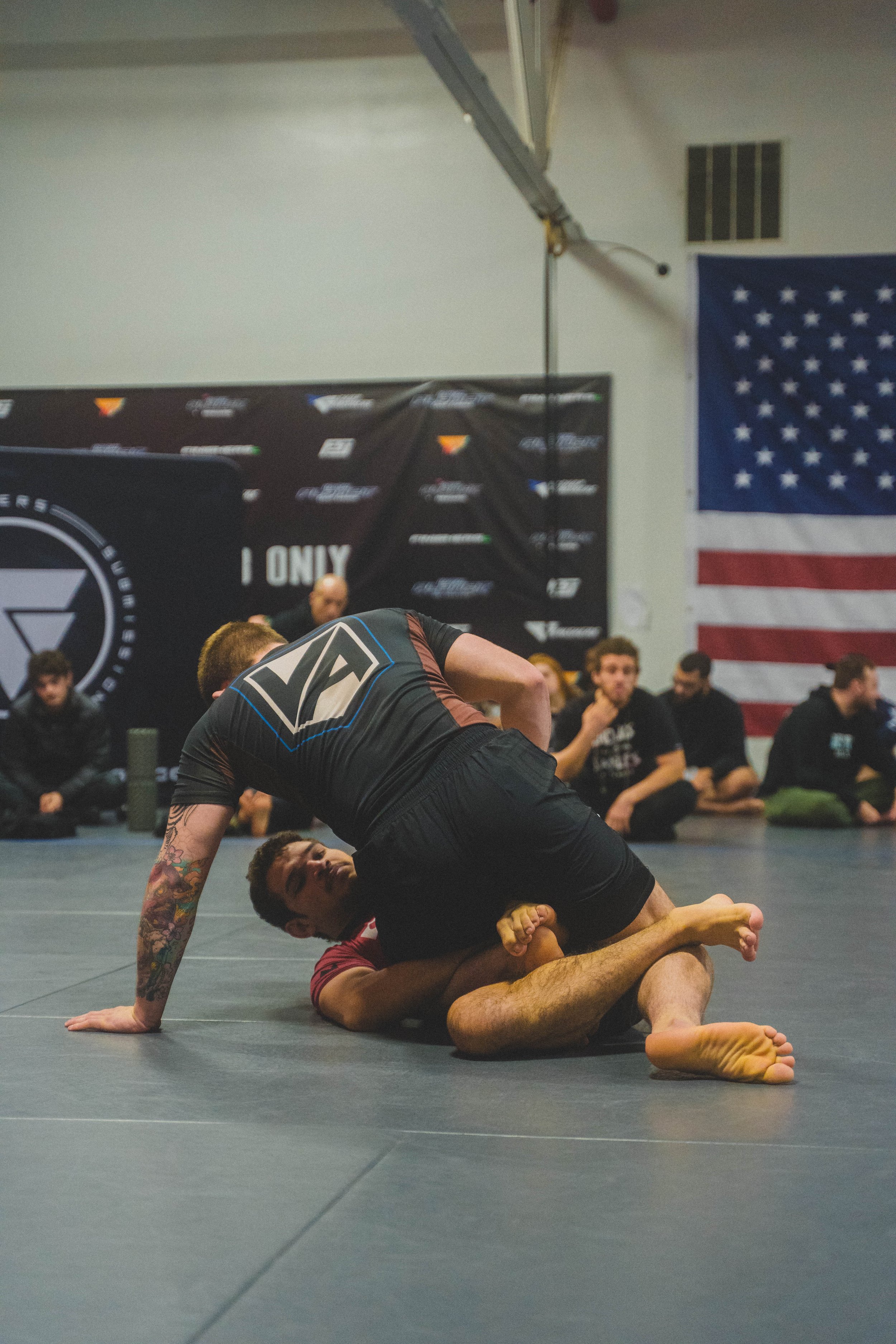 Two male fighters in mixed martial arts match; one in black shorts is on top, pinning the other in maroon shirt to the mat during a grappling exchange in a gym with American flag and sponsorship banners in background.