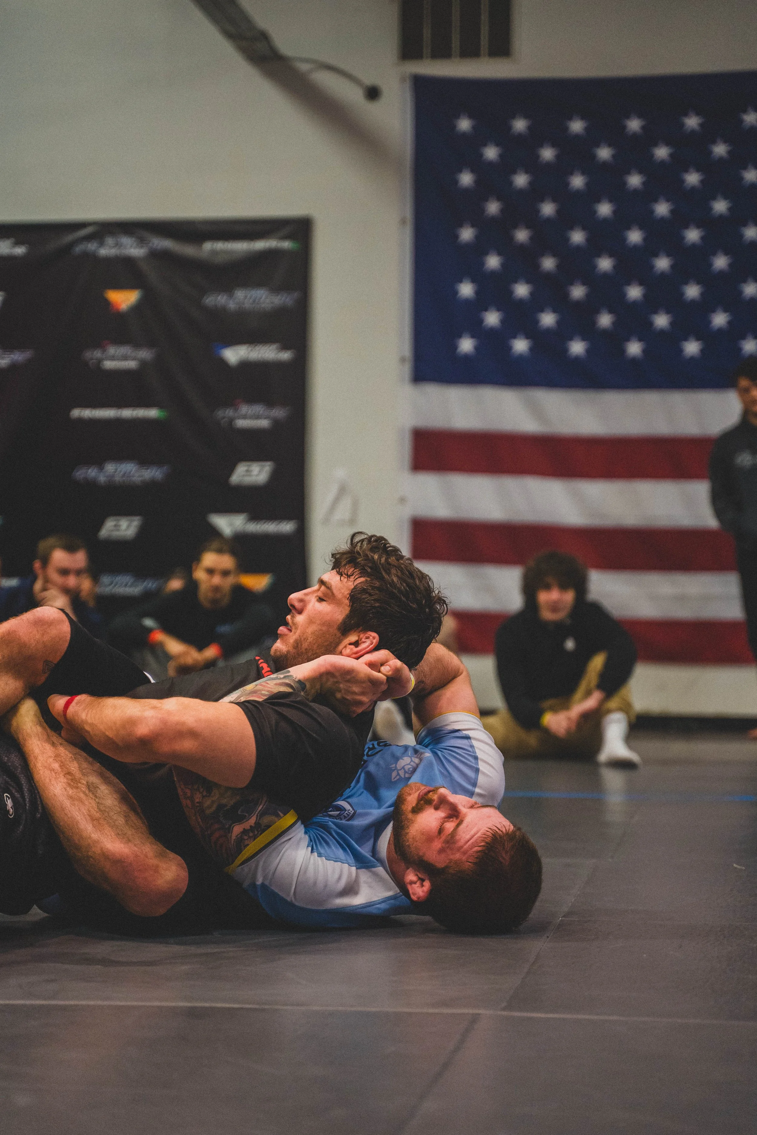 Two men grappling on the floor during a wrestling match, with several spectators and American flags in the background.