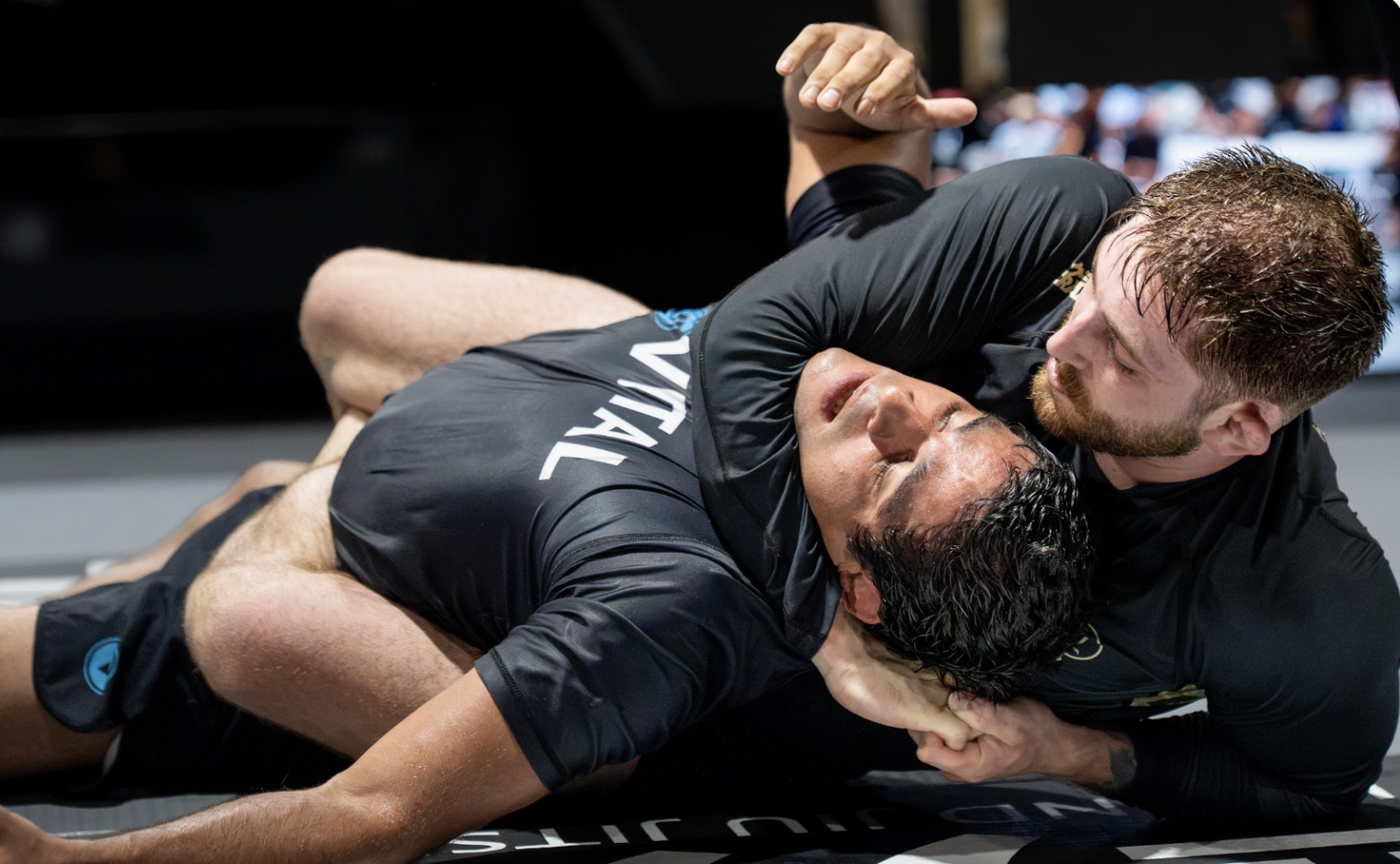 Two male mixed martial artists, one in a black shirt, grappling on the mat during a fight.