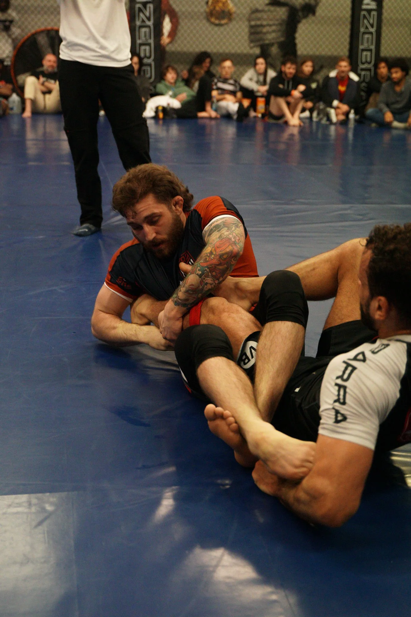 Dylan grappling on a blue wrestling mat during a Brazilian Jiu-Jitsu match, with an audience watching in the background.