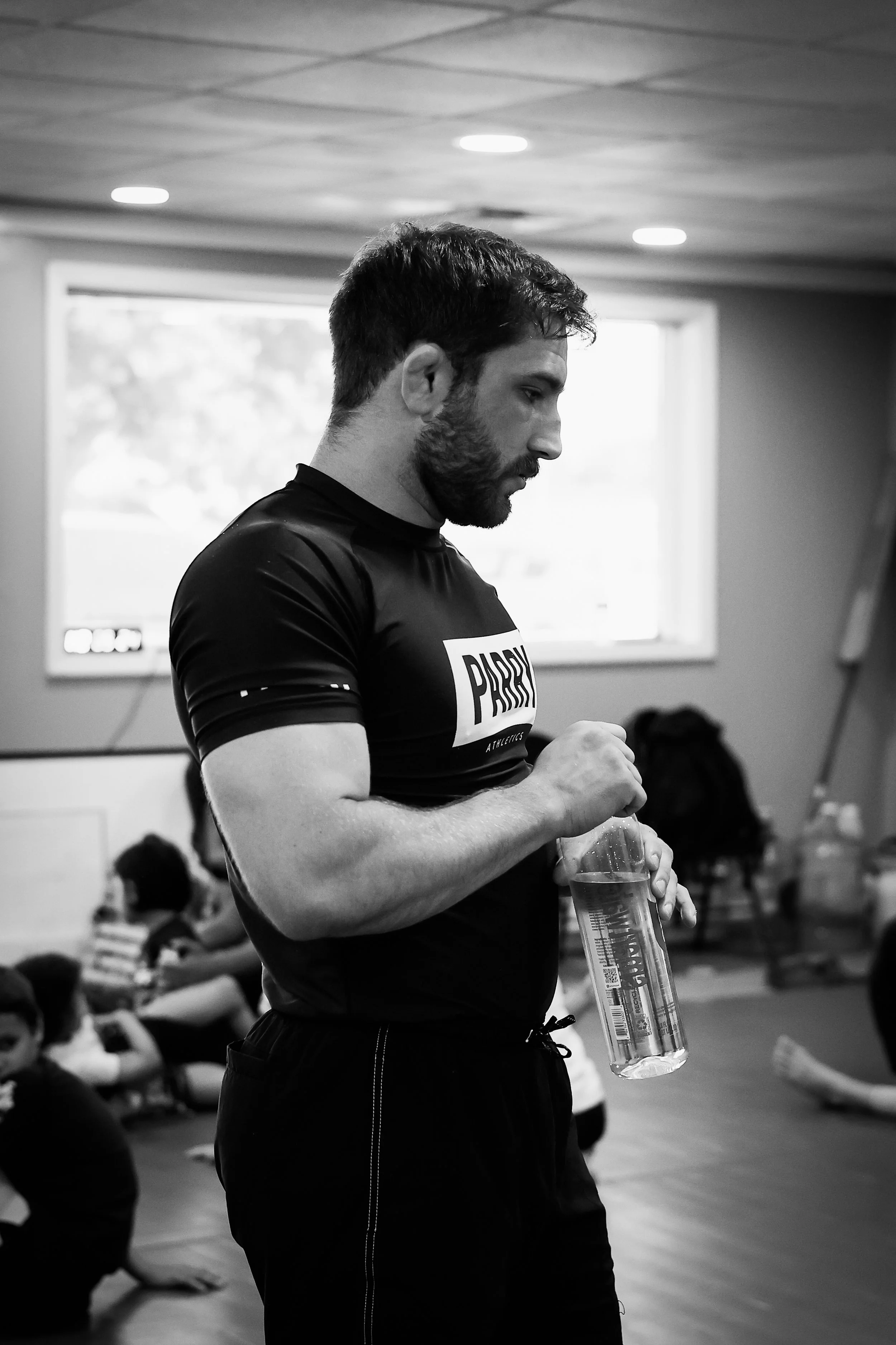 A man with a beard wearing a black athletic shirt holding a large water bottle in a room with children and windows in the background.