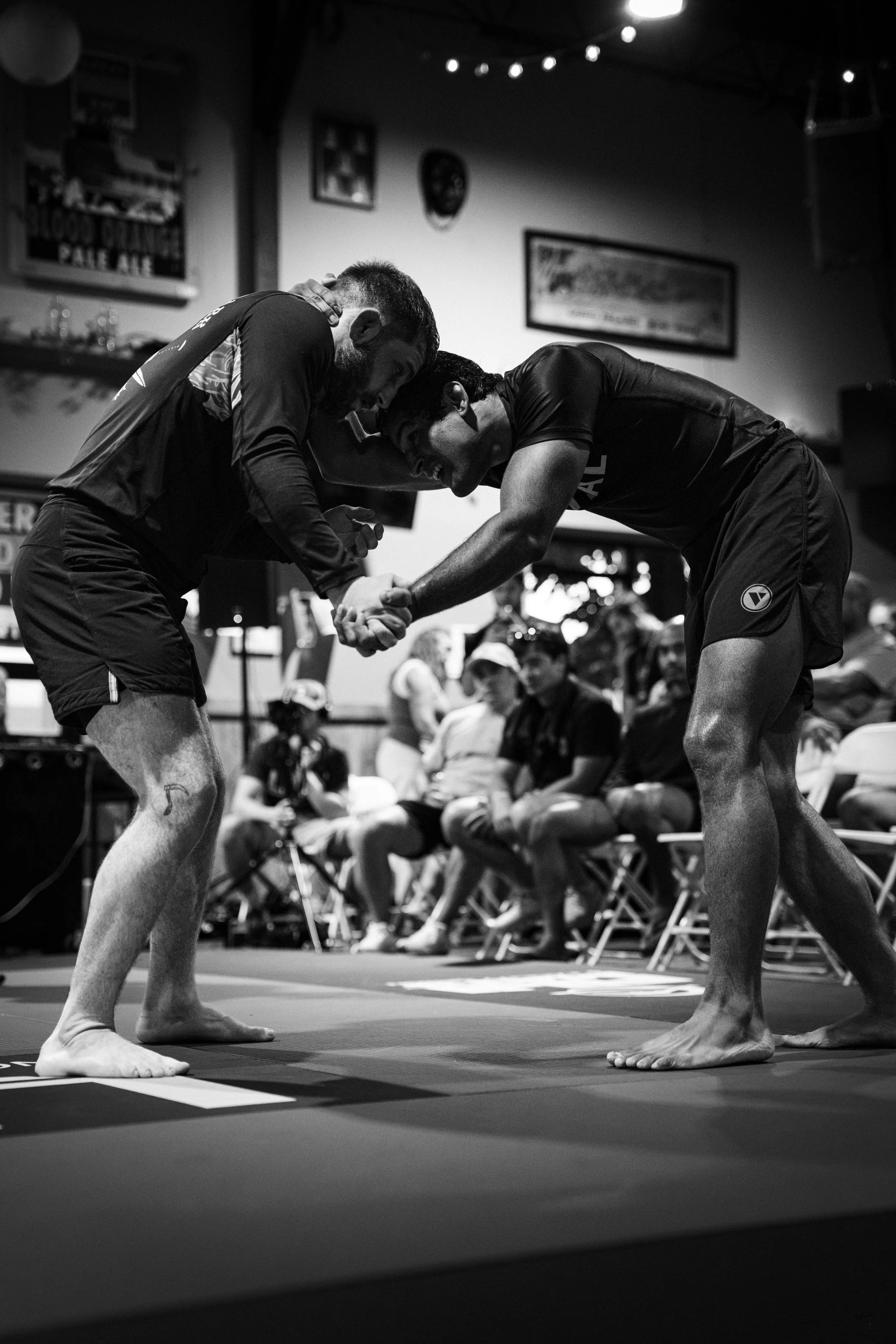Two men engaged in a grappling match in a martial arts competition, holding each other's wrists and leaning in on a mat, with spectators seated in the background.