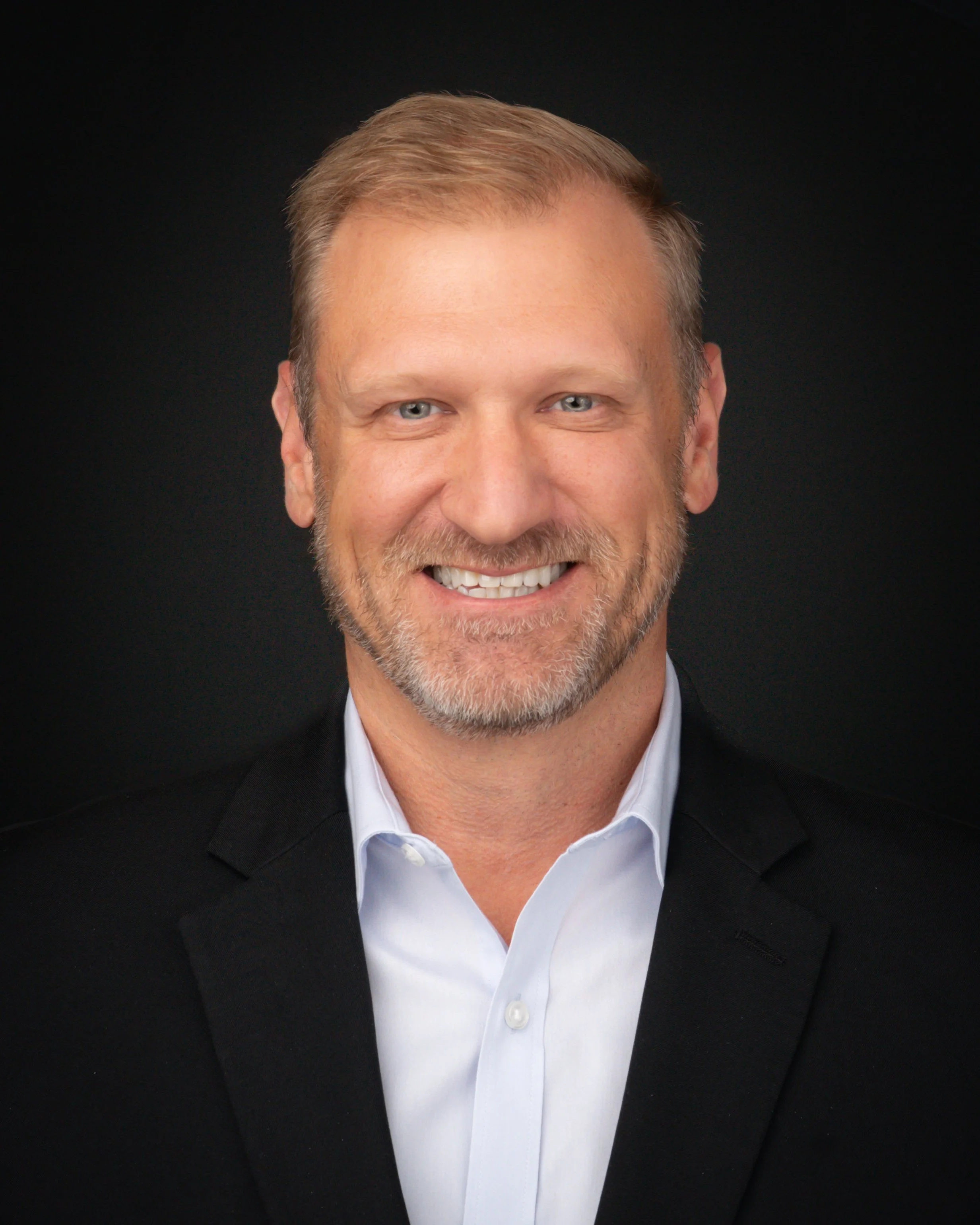 Close-up of a middle-aged man with light brown hair and beard wearing a white shirt and black blazer, smiling against a black background.