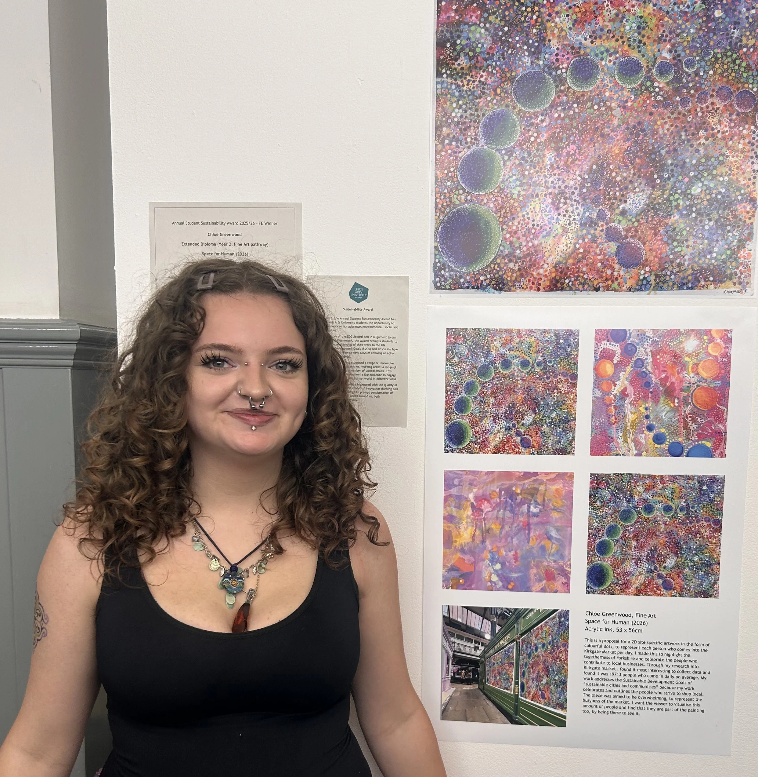 A woman with curly hair, wearing a black top and layered necklaces, smiling in front of a colorful art display on a gallery wall. The display includes vibrant, abstract paintings with circular patterns and a photograph of an outdoor scene with a store front.
