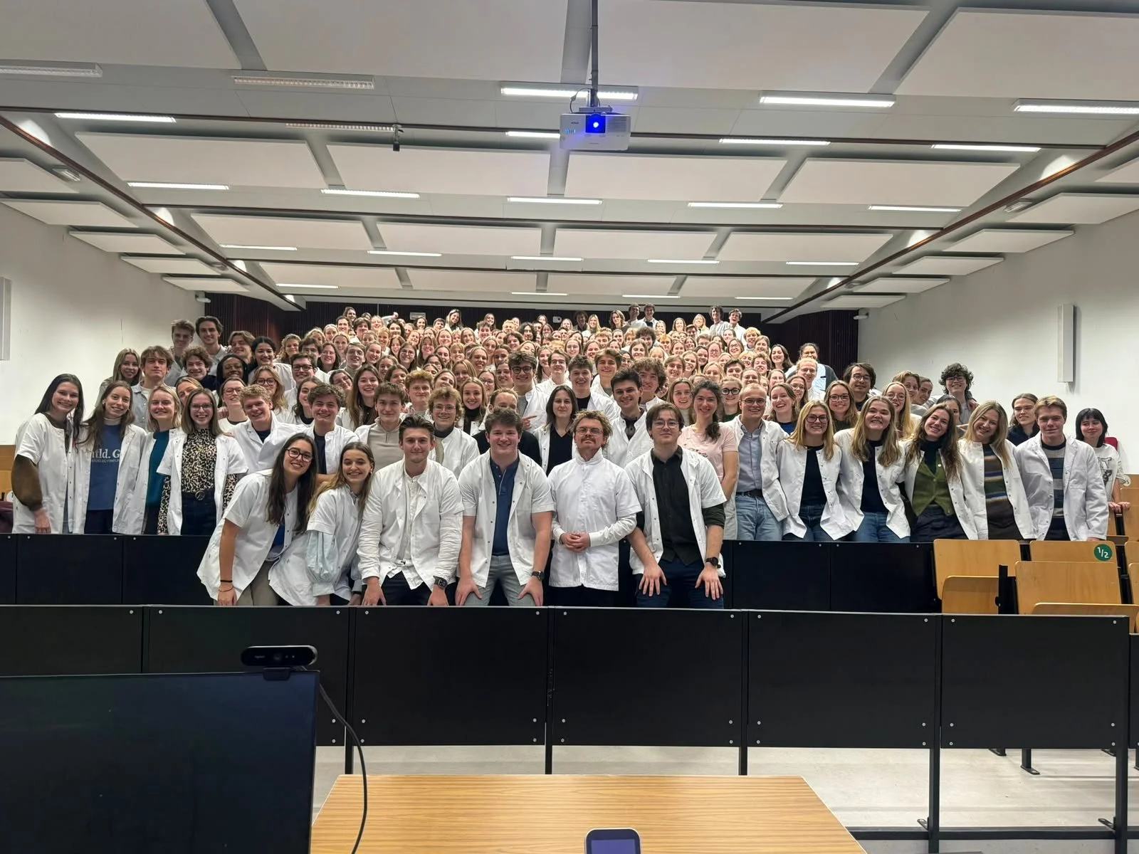 Grote groep mensen, voornamelijk jonge volwassenen, poseert in een auditorium op een groepsfoto. Ze dragen witte labjassen en lachen naar de camera.