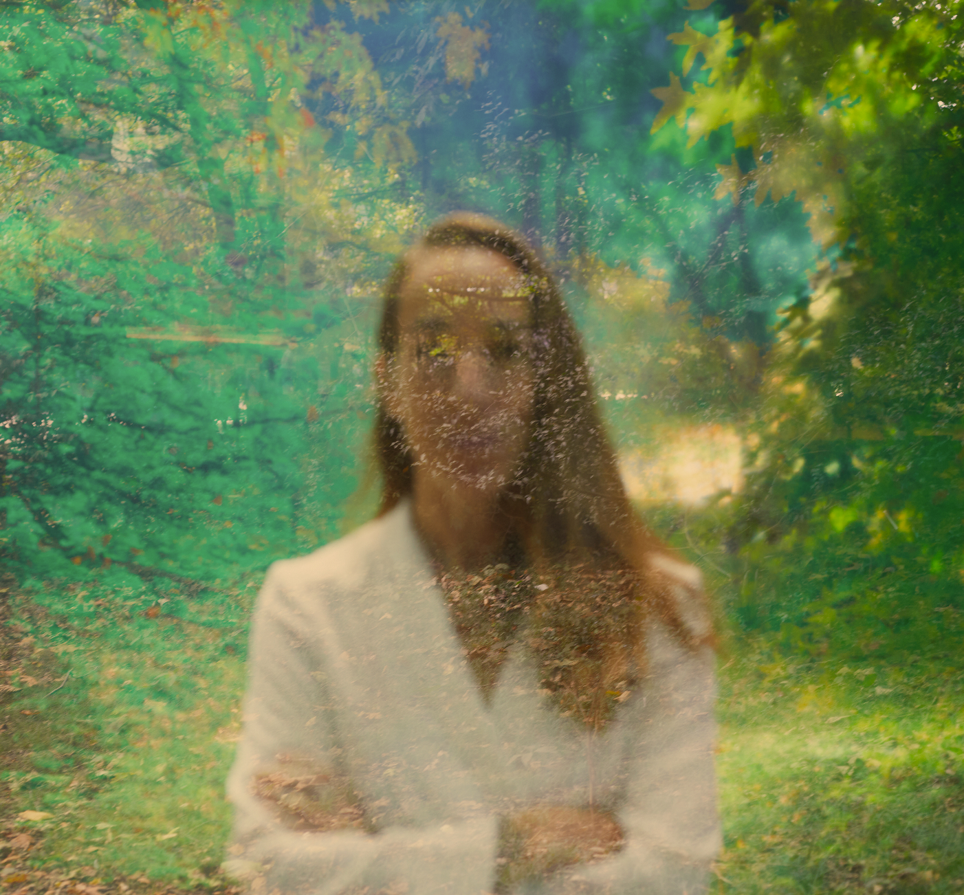 A person with long hair and glasses standing in front of a green, foliage-filled background, with a reflection of trees and a girl in a glass window or door.