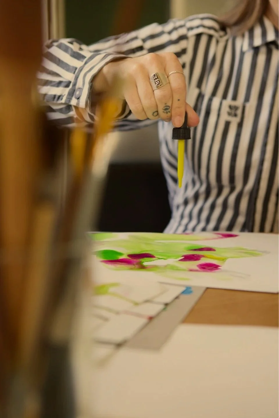 Person wearing a striped shirt holding a dropper with yellow liquid over a colorful abstract painting.