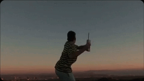 Person taking a selfie outdoors at sunset with a cityscape in the background.