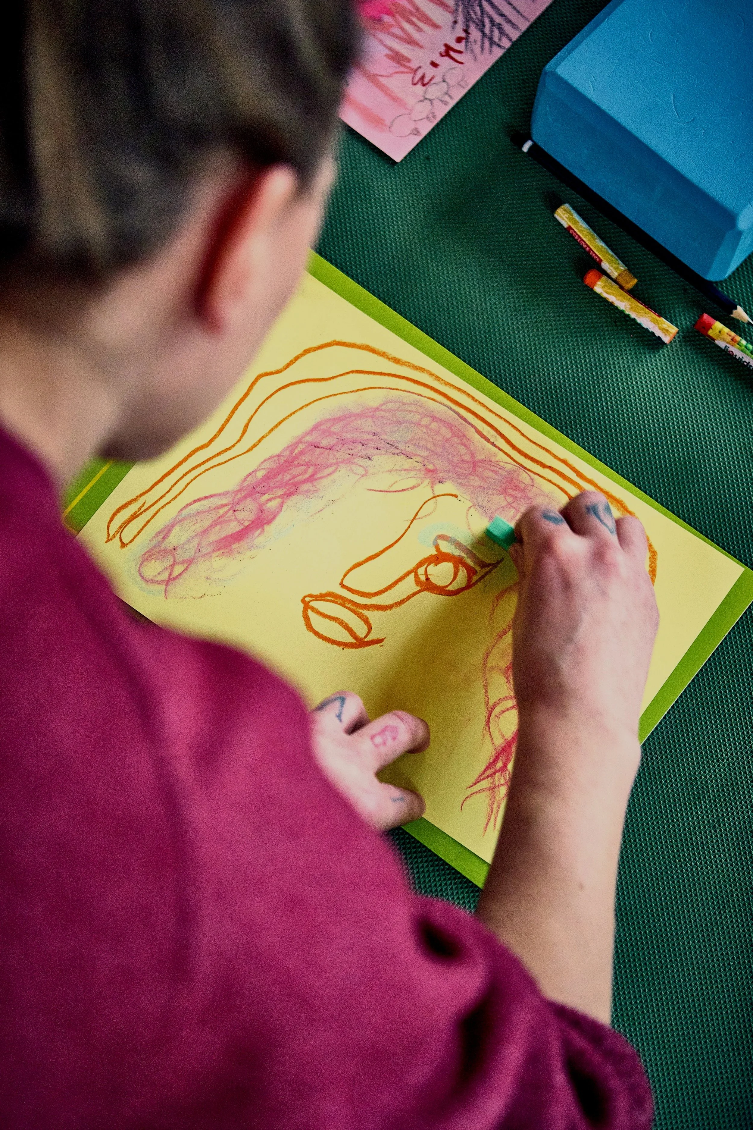 Child drawing a rainbow on yellow paper with orange and pink crayons, wearing a pink shirt, on a green textured surface, with other art supplies visible.