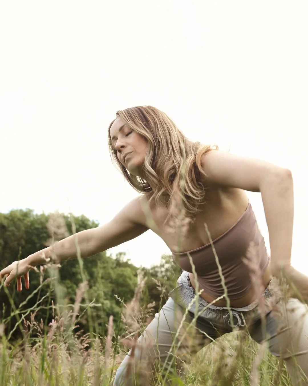Woman with brown hair wearing a strapless top and loose pants in a grassy field during daytime.