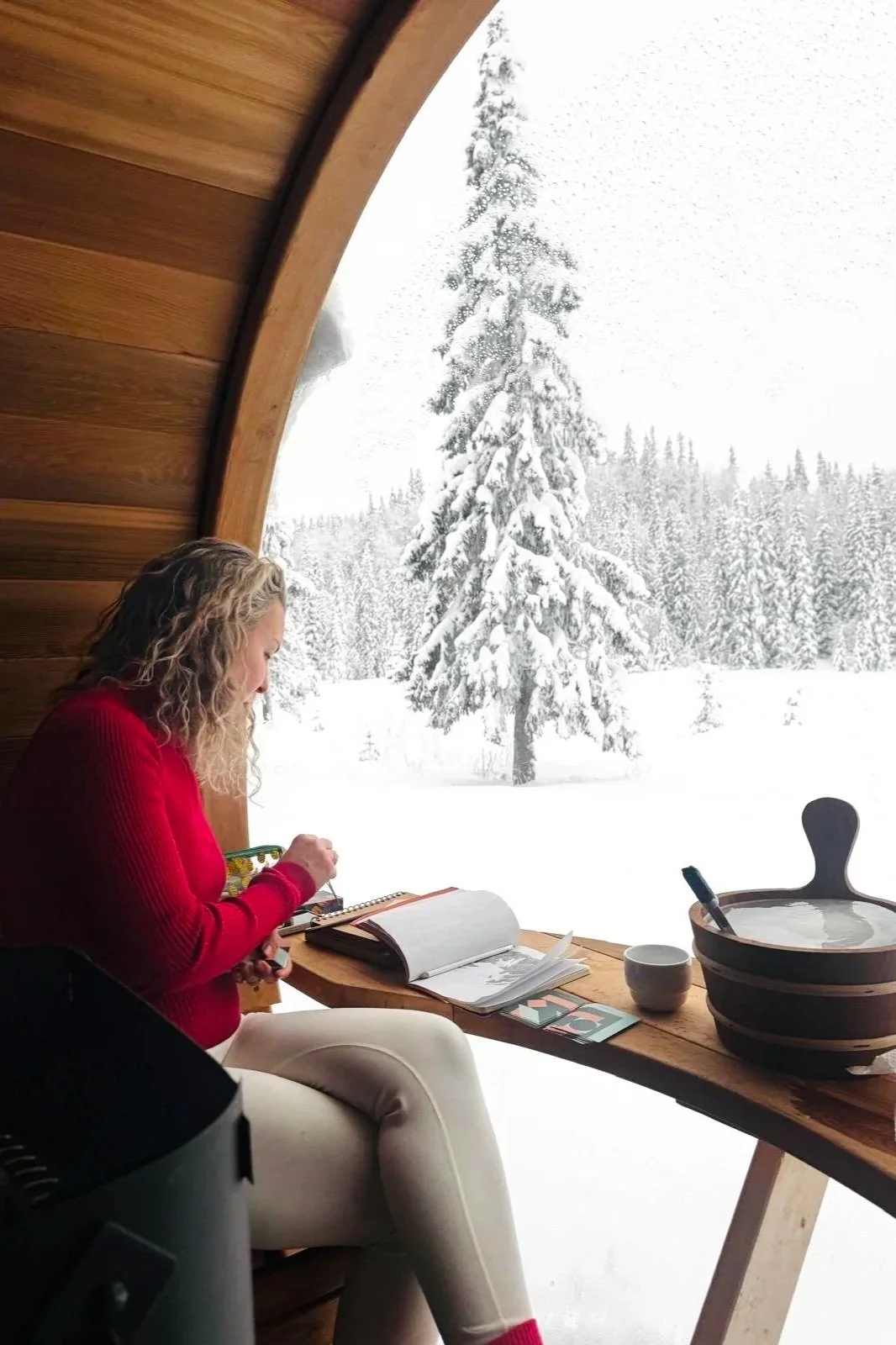 A woman with curly blonde hair wearing a red sweater sitting by a large window in a wooden cabin, with snow-covered trees outside.