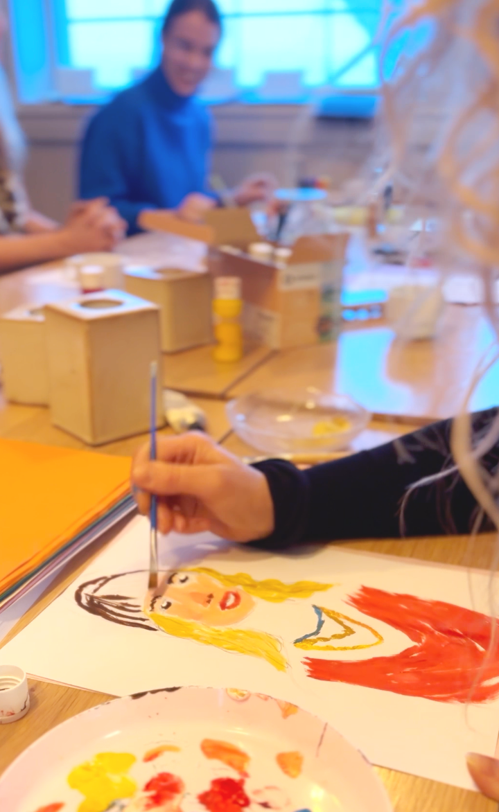 A person painting a portrait of a blonde woman wearing a red shirt on paper, with art supplies and other people working in the background.