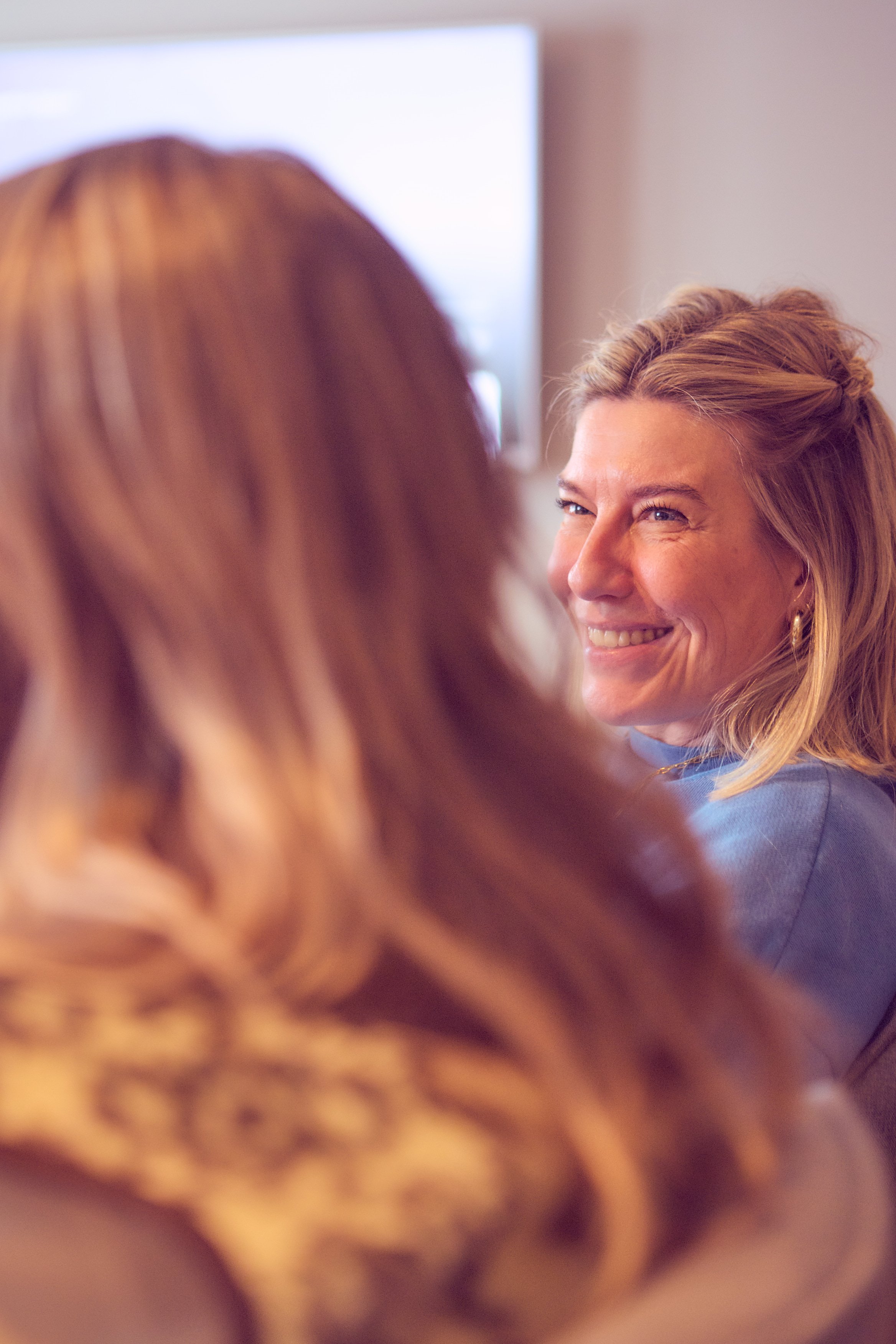 Smiling woman with light brown hair styled in loose waves and a braid, wearing a blue top, engaging in conversation with another person with reddish hair, indoors with a neutral background.