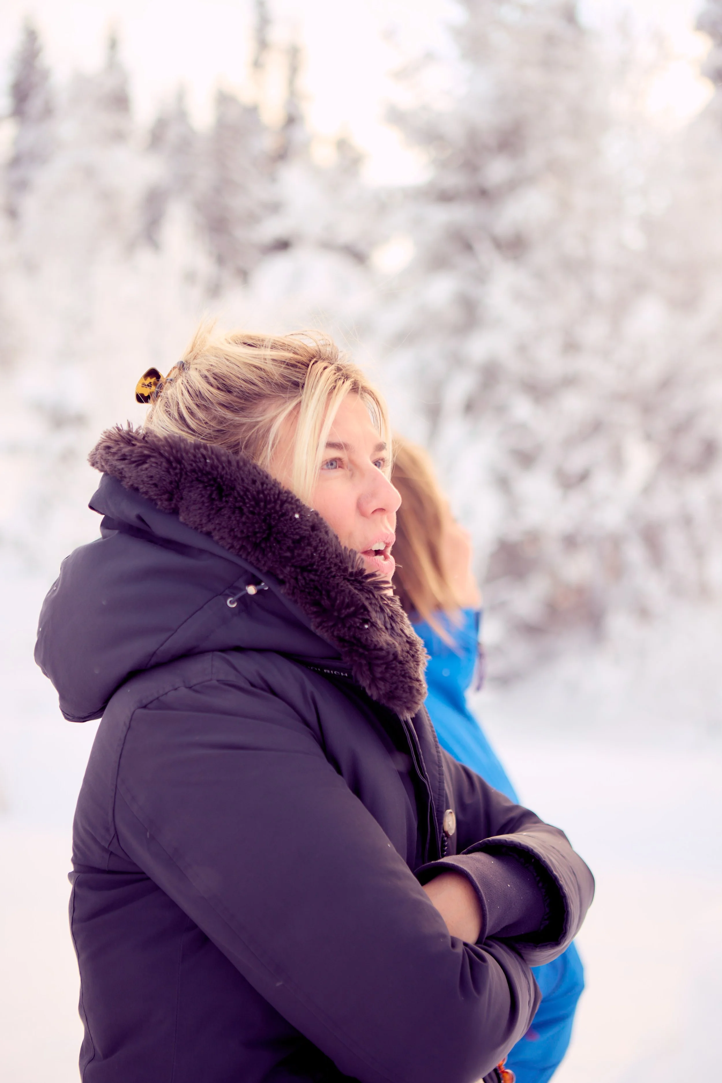 A woman in a black winter coat with a fur-lined hood stands outside in a snowy landscape during daytime, with her arms crossed and looking to the right.