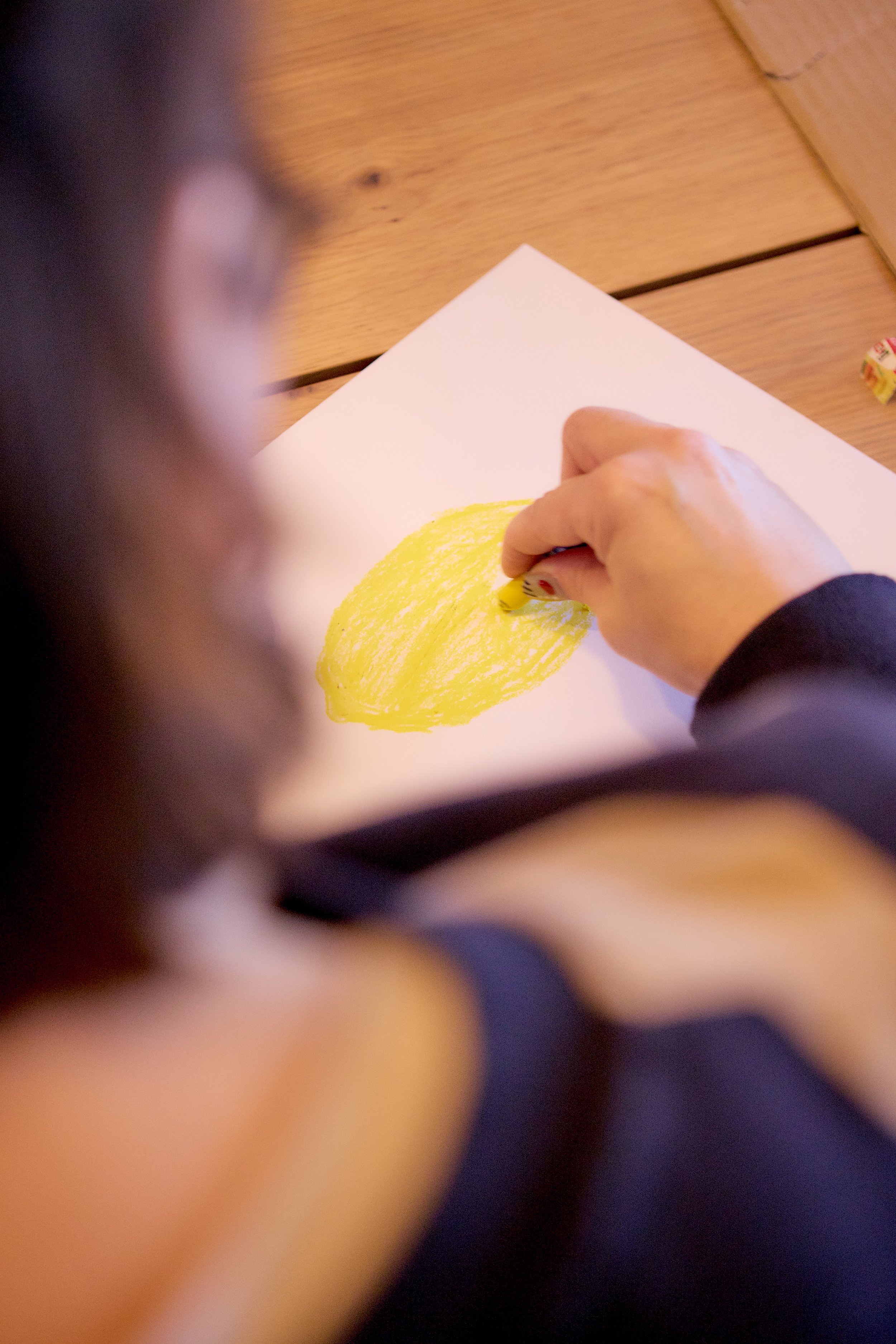 A person's hand holding a yellow crayon and drawing on a white sheet of paper. The drawing appears to be a large yellow shape.
