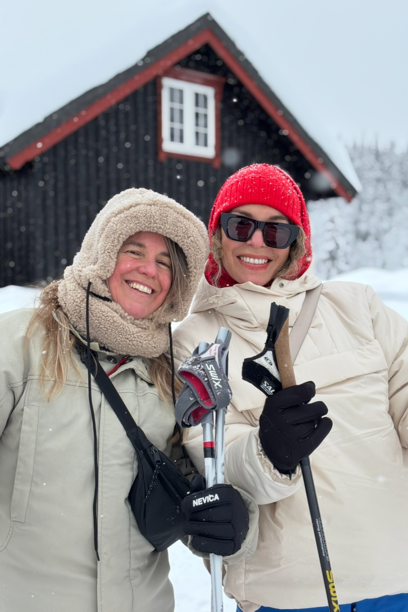 Two women in winter clothing standing in a snowy landscape, smiling at the camera, with a dark wooden house with red trim in the background. One woman is wearing a beige hooded coat and scarf, and the other is wearing a white coat, red knit hat, and 