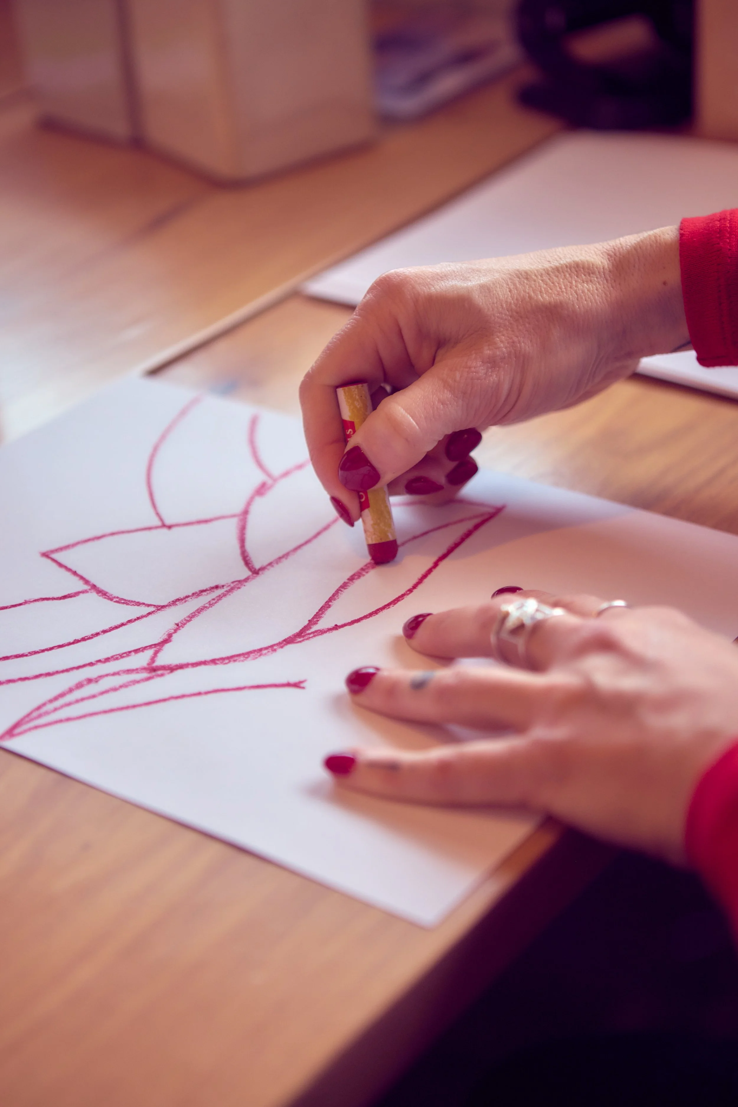 A person with red painted nails is drawing on a piece of paper with a red crayon. The drawing appears to be of a large leaf or flower. The scene is set on a wooden table with some papers and office supplies in the background.