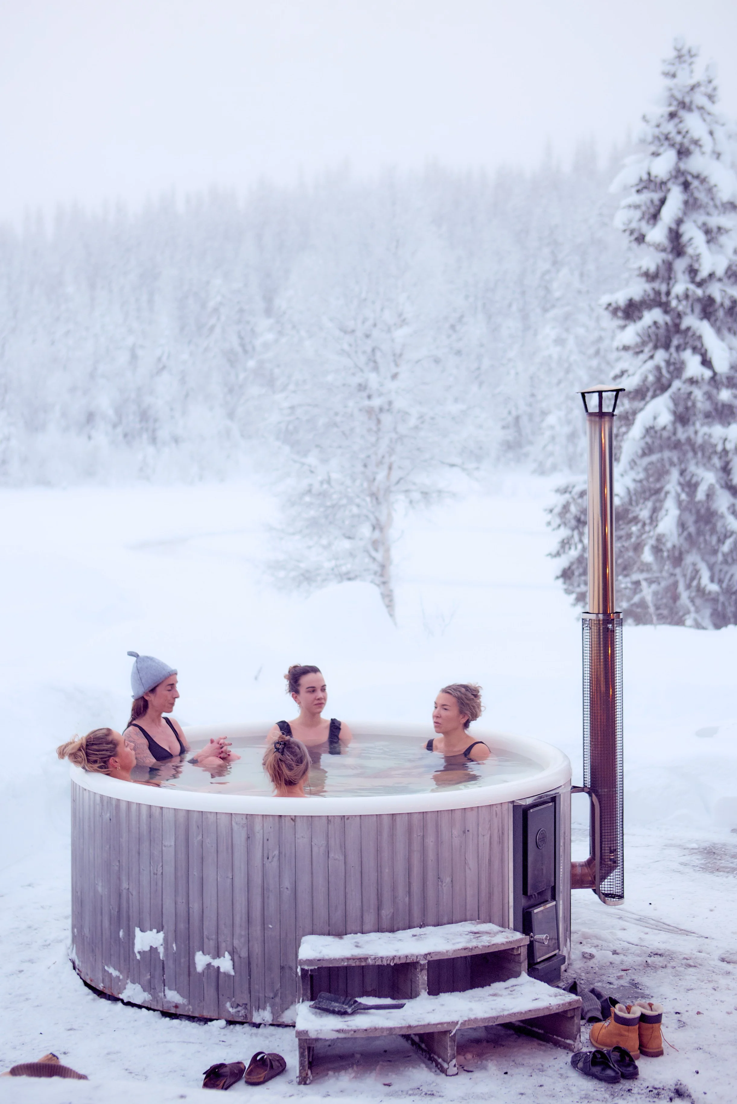 Group of women relaxing in an outdoor hot tub in a snowy landscape surrounded by snow-covered trees with a chimney emitting heat nearby.
