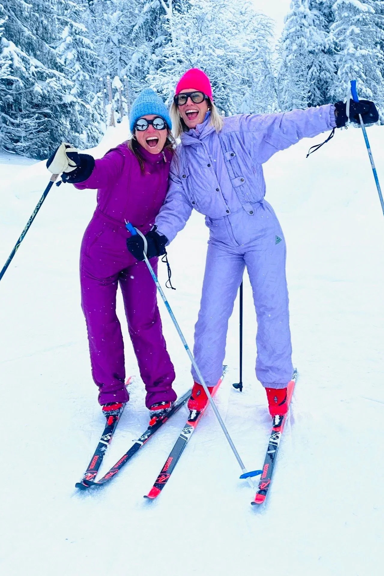 Two women skiing in a snowy forest, wearing colorful winter outfits and sunglasses, smiling and posing for the photo.