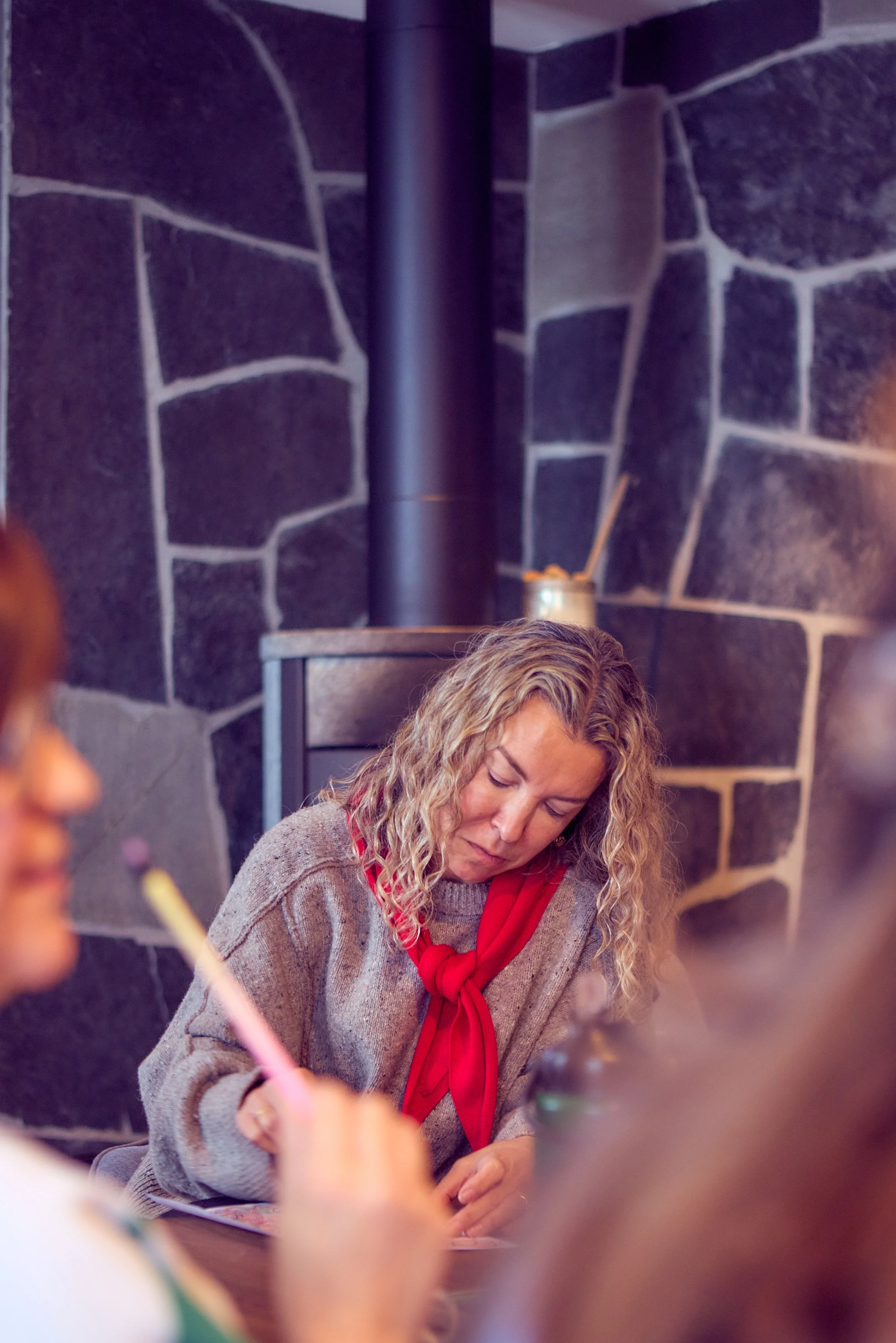 A woman with curly blonde hair wearing a gray sweater and red scarf, sitting at a table in a room with dark stone walls and a black cylindrical chimney, looking down.