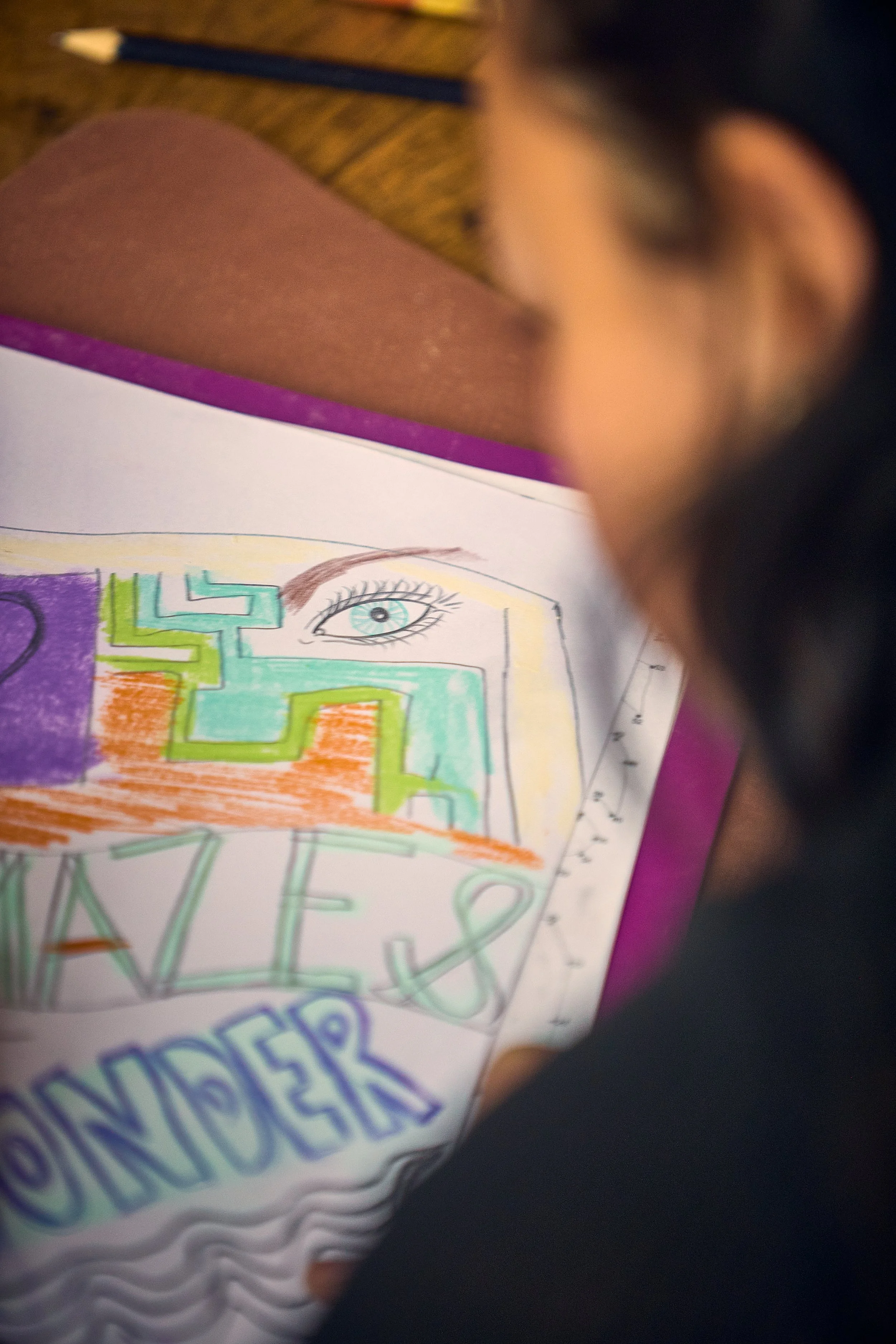 A person with glasses is coloring a paper with a drawing and the words 'HAPPY HOLIDAYS' written on it. The drawing features an eye and colorful geometric shapes.