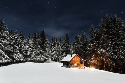 A snowy landscape at night with a small wooden cabin illuminated from inside, surrounded by snow-covered trees under a starry sky.
