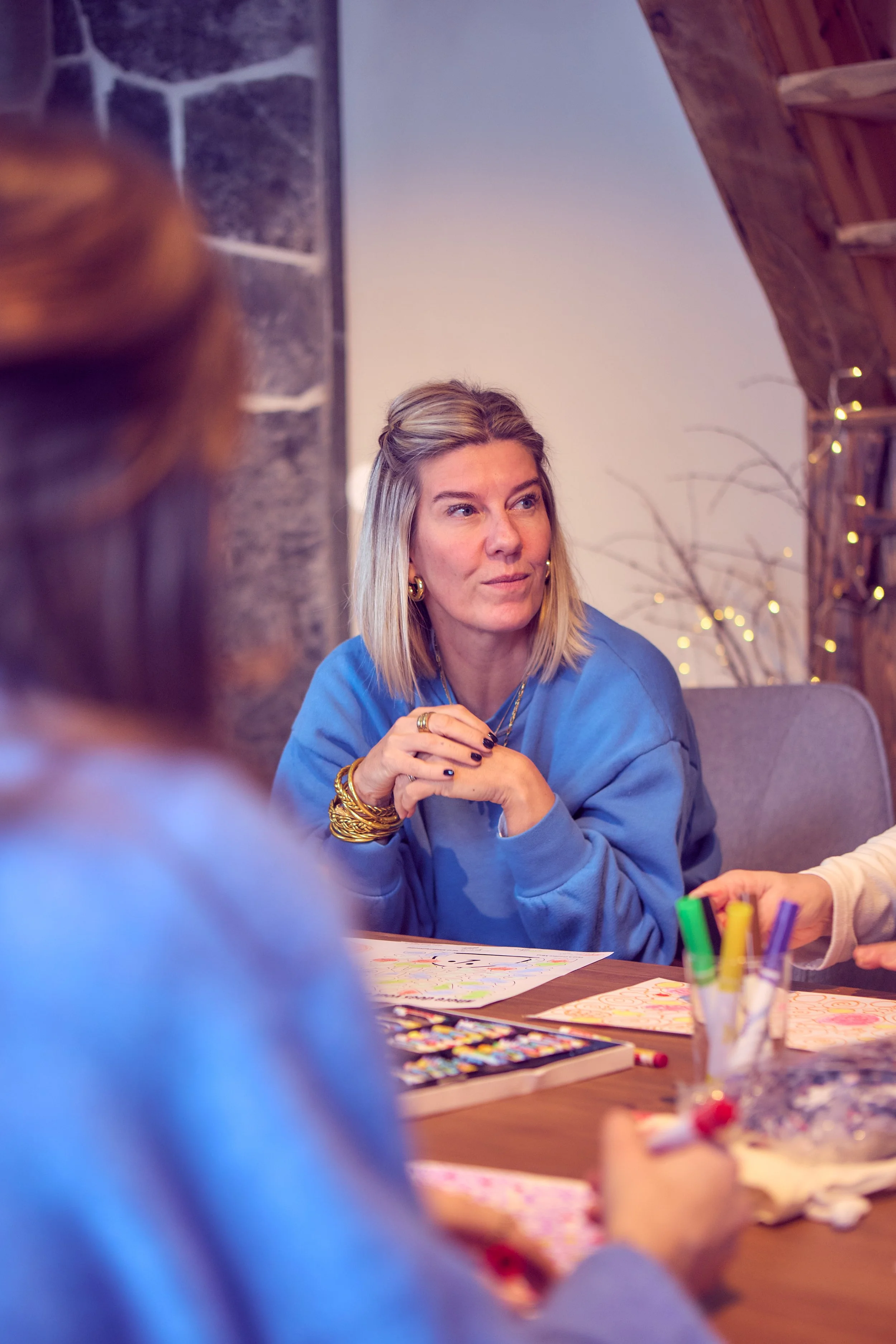 A woman with blonde hair and wearing a blue sweatshirt sitting at a table, engaged in conversation with children during a crafting activity. The table has coloring sheets, markers, and art supplies. The background features a wall with string lights and decorative branches.