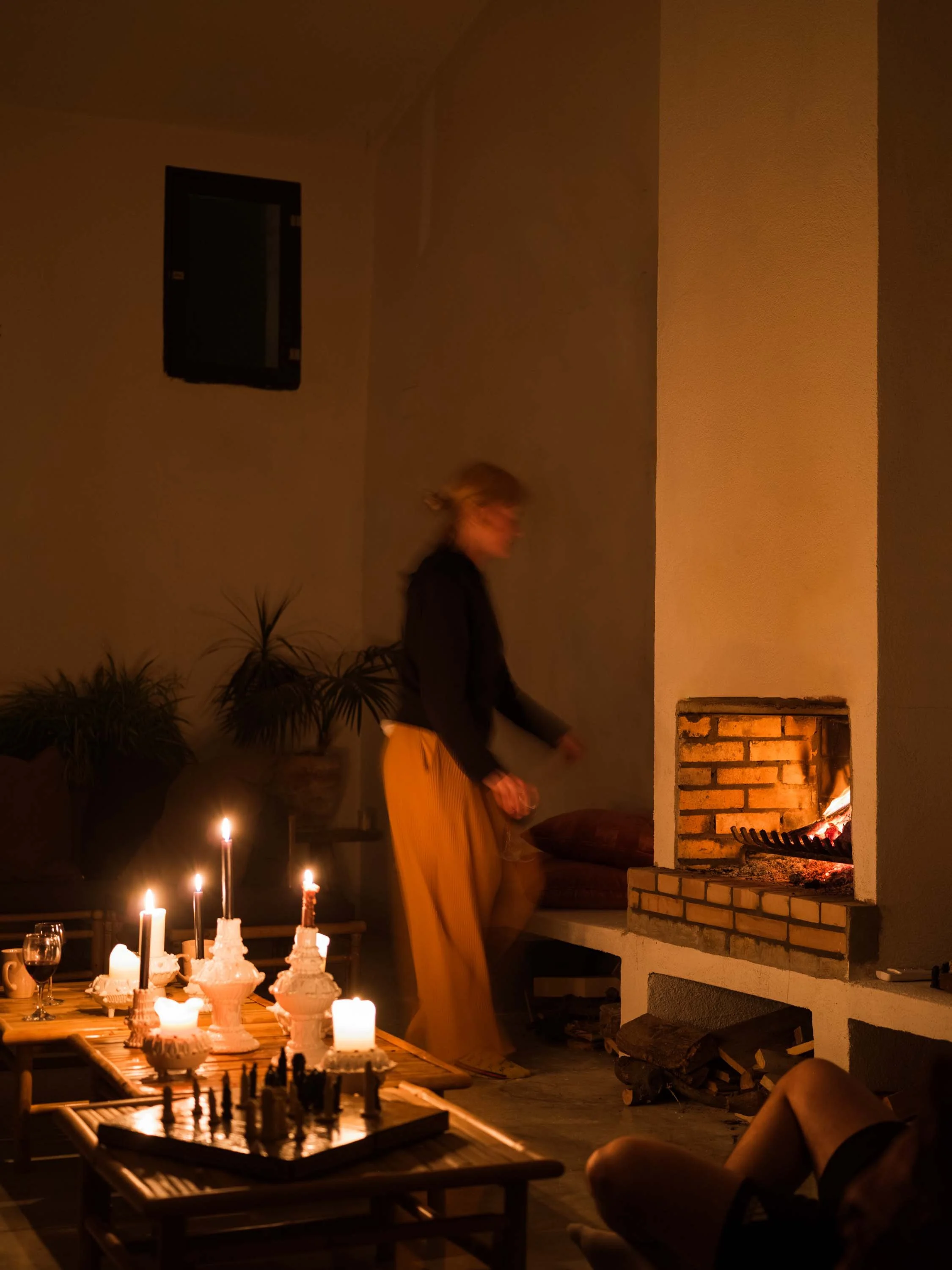 A person in a yellow skirt and black top standing near a fireplace with burning firewood. There are candles on the table in the foreground and a chessboard. The setting appears to be a cozy indoor space during evening or night.