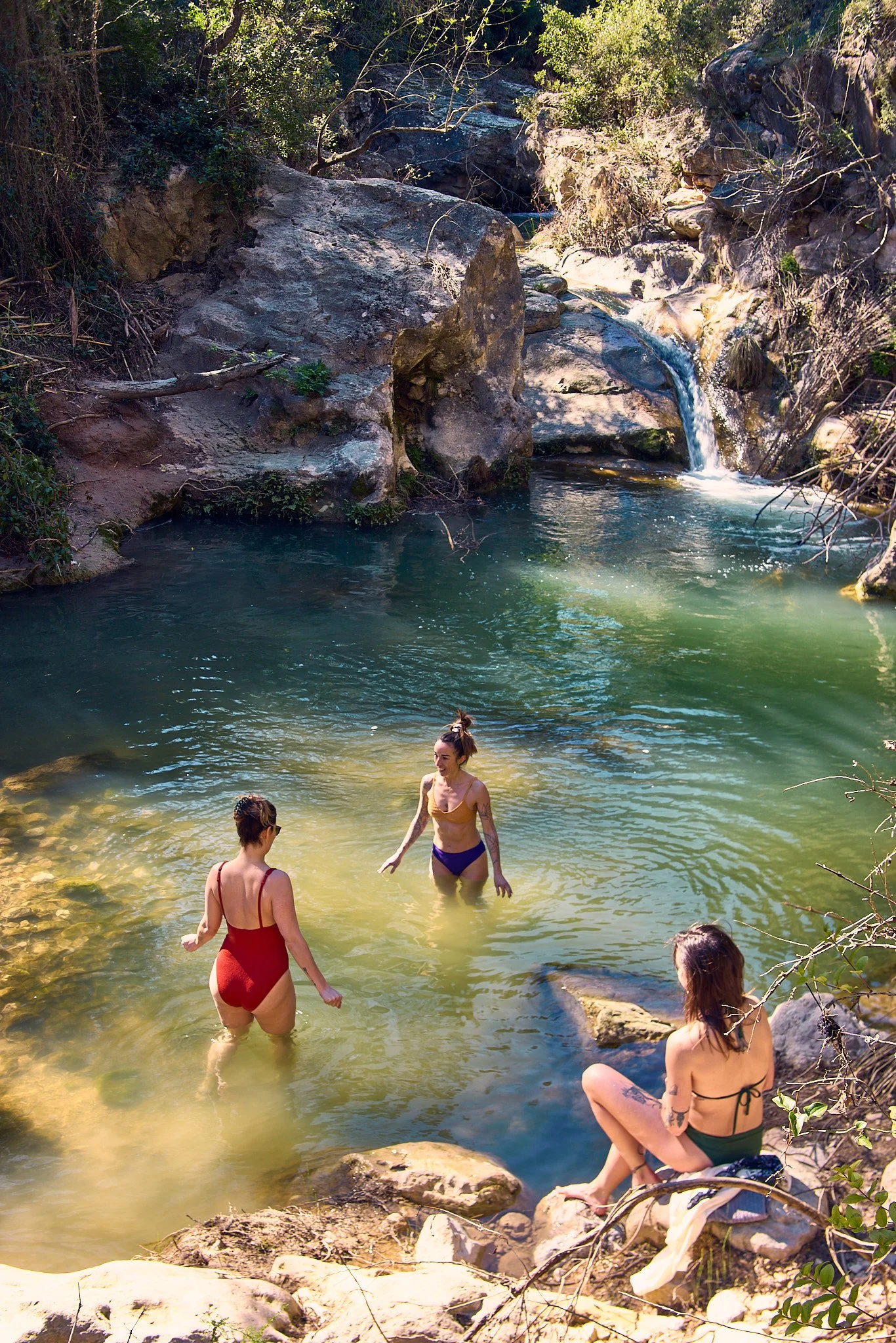 People in swimsuits enjoying a natural waterfall and swimming hole surrounded by rocks and trees.