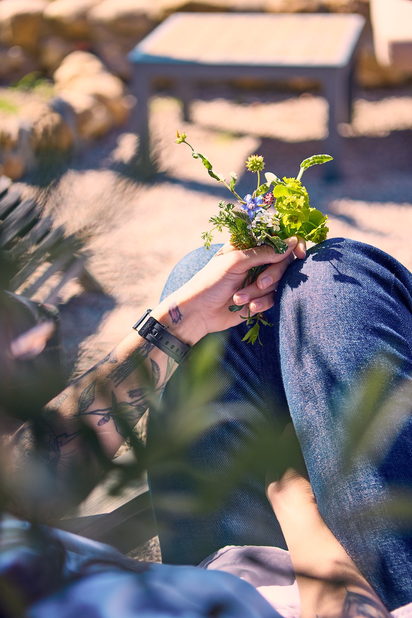Person sitting outdoors holding a small bouquet of mixed flowers, wearing blue jeans and a tattooed arm with a black wristwatch, in a setting with sunlight, rocks, and blurred background.