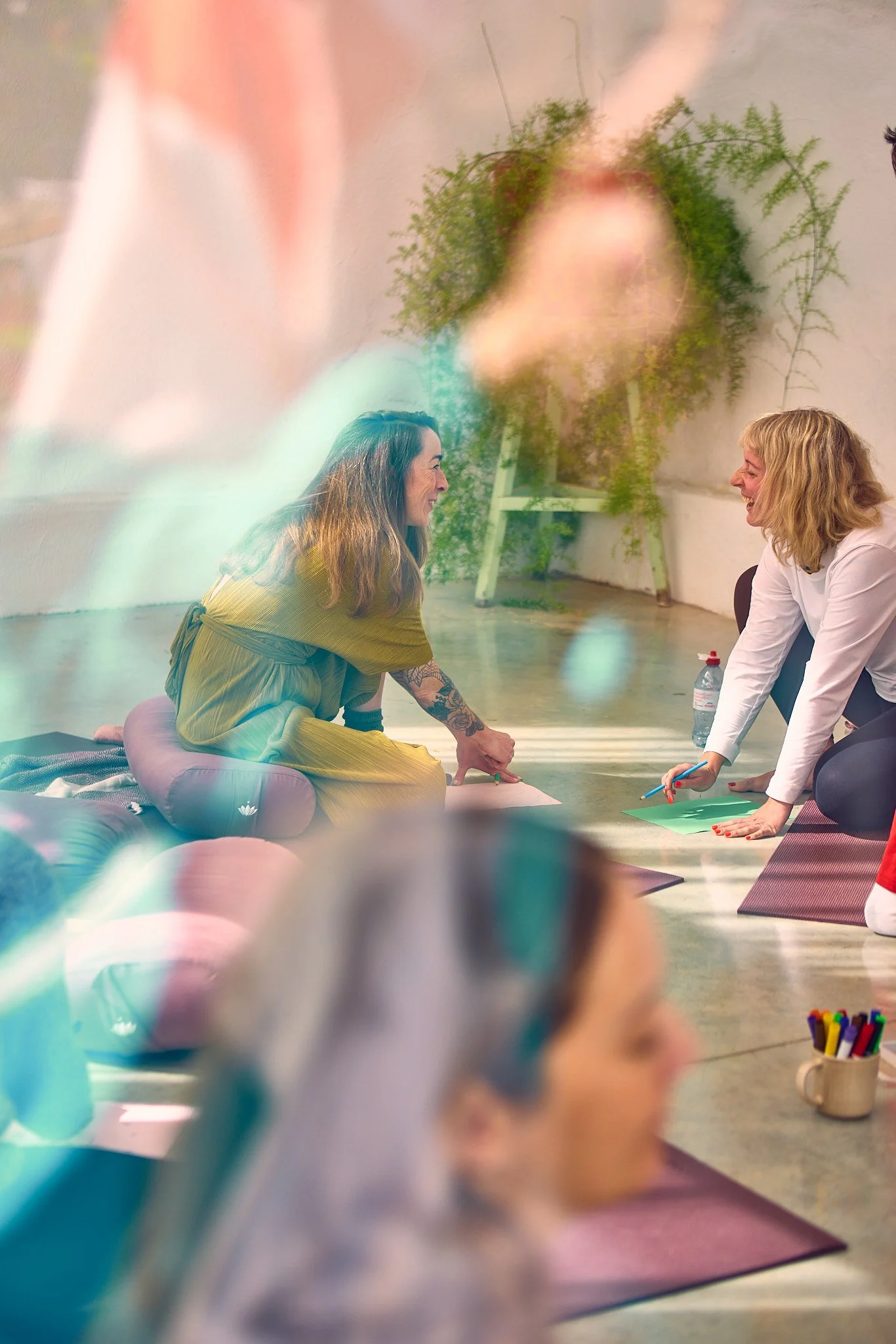Two women are smiling and sitting on exercise mats, engaging in a fun workshop or class, with various art supplies and a potted plant in the background.