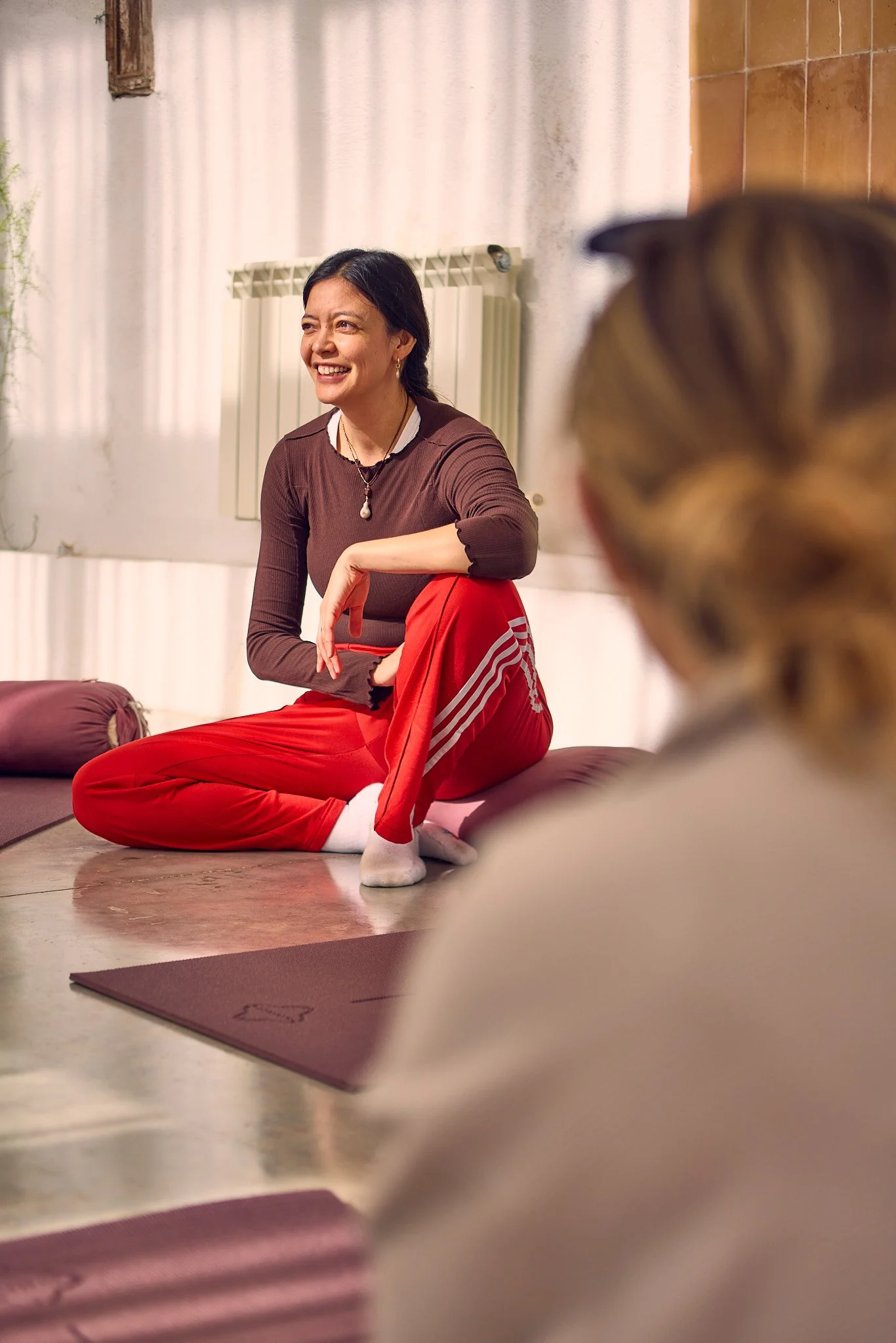 A woman sitting on a yoga mat, smiling, with another person in the foreground, in a room with a white wall and a radiator.