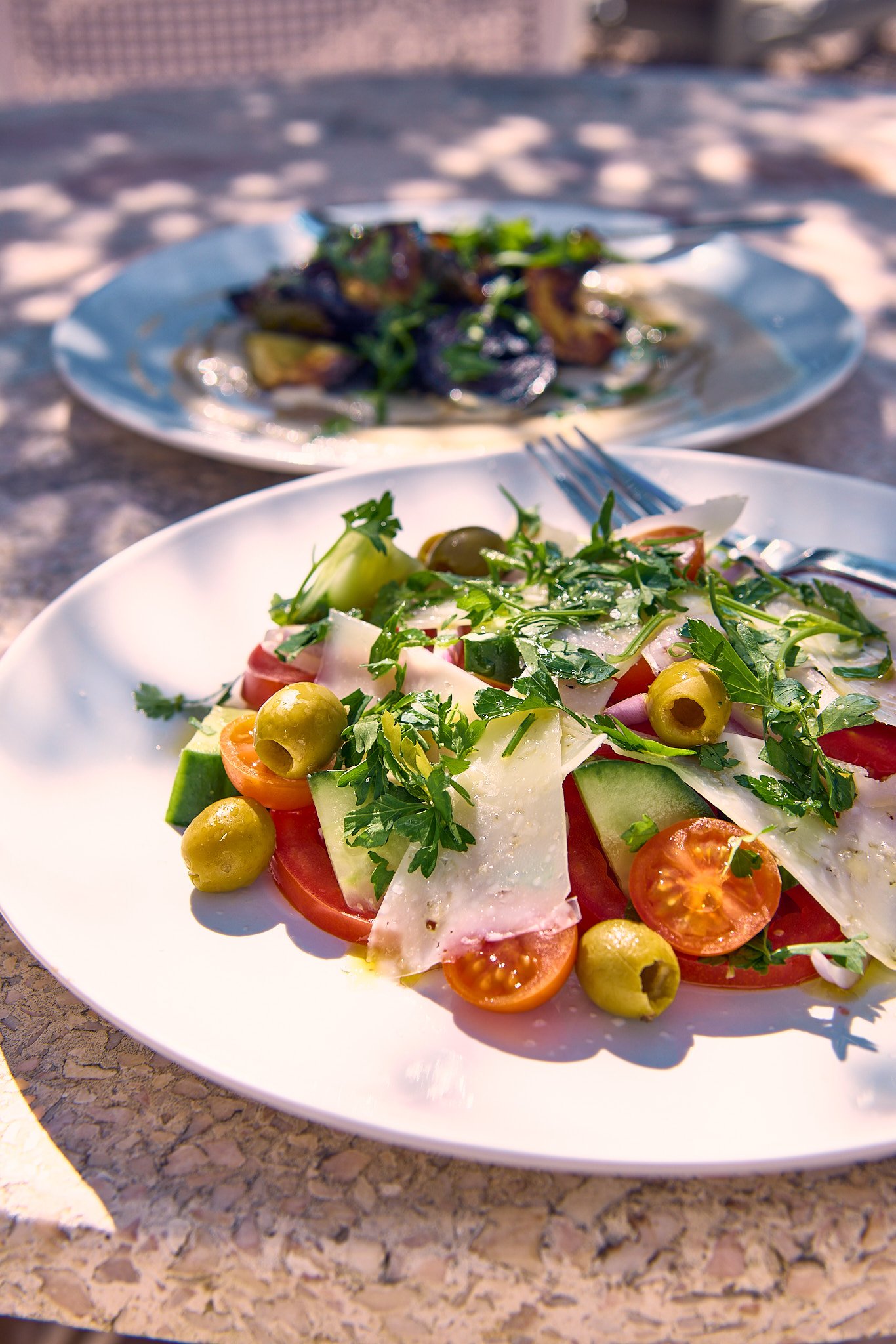 A colorful salad with tomatoes, green olives, cucumbers, sliced meats, and fresh greens on a white plate, with another dish of cooked food in the background, on a textured outdoor tabletop.