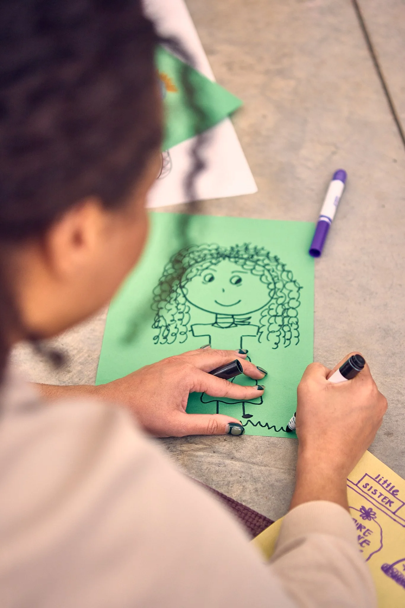 Child coloring with a black marker on green paper, drawing a girl with curly hair, wearing a striped shirt and skirt. There are other papers and markers on the table.