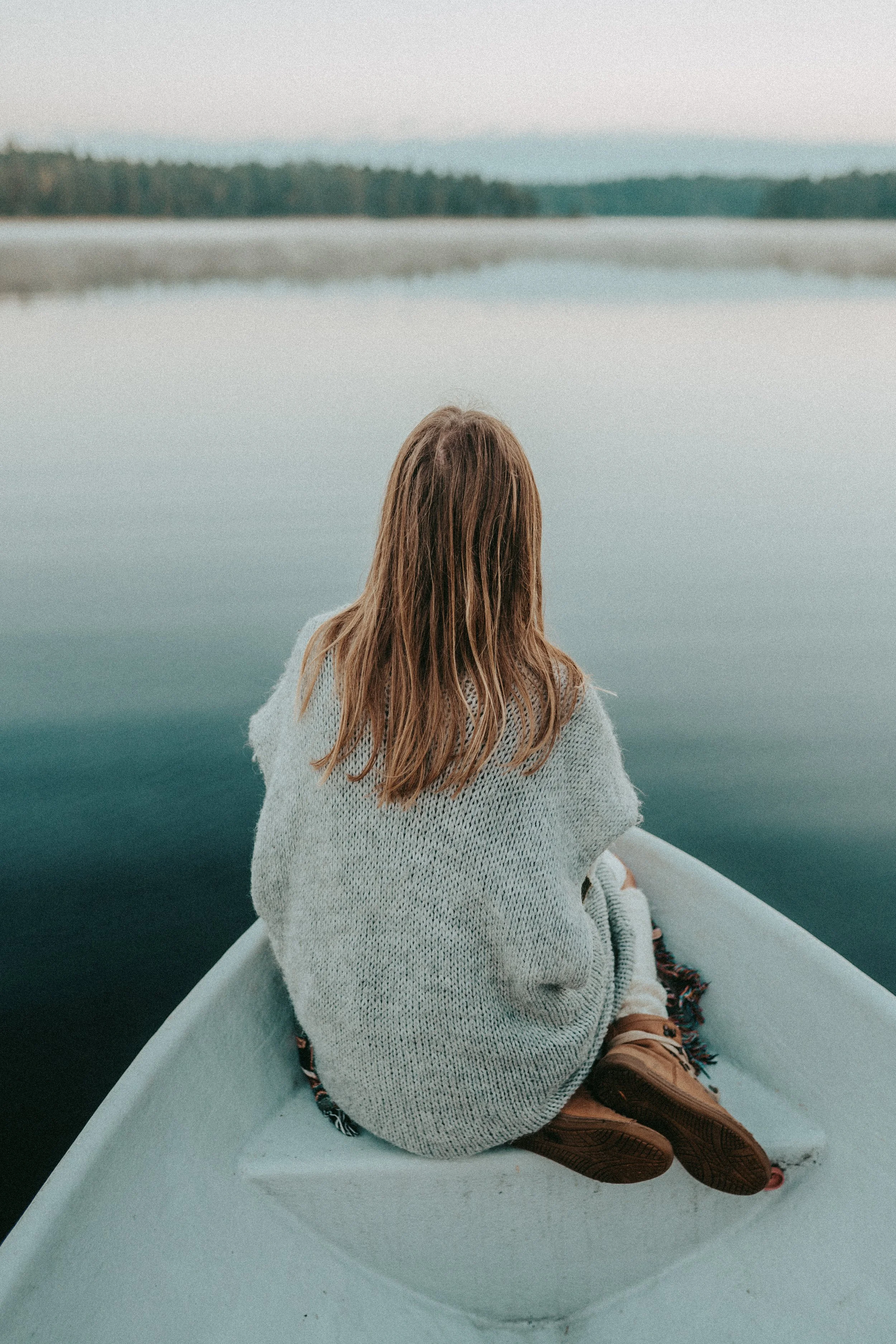 A person with long brown hair sitting in a small boat on a calm body of water, wearing a light-colored sweater and brown shoes, facing away from the camera with a distant tree-lined shoreline in the background.