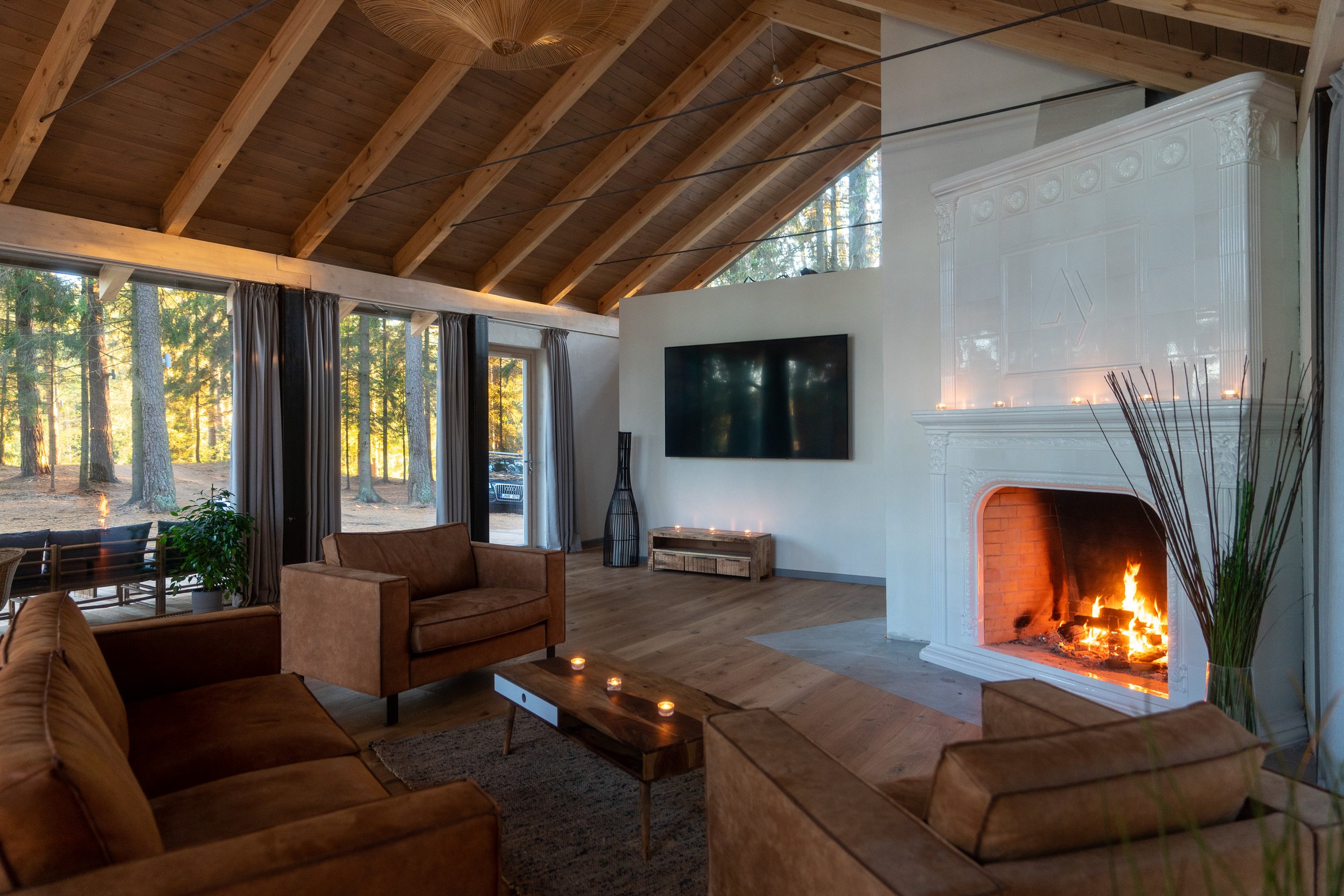 Interior of a modern living room with a high wooden vaulted ceiling, large windows with curtains, a fireplace with a fire, and comfortable seating.