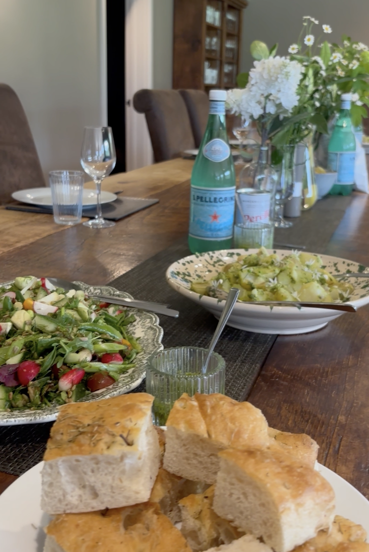 A dining table set with bread, salad, and bottled drinks, with a vase of white flowers in the background.