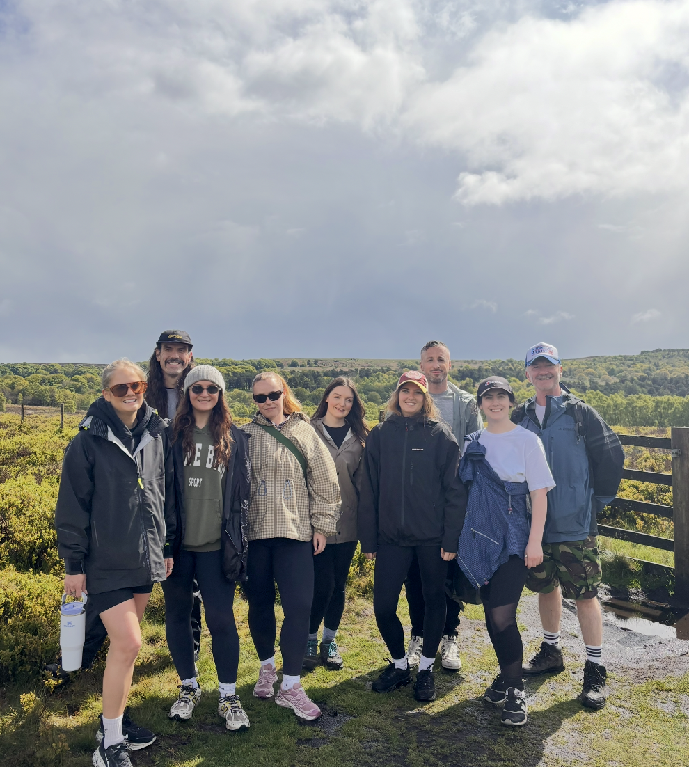 Group of nine people, mostly young women and two men, standing outdoors on a hiking trail in a rural area with green fields and hills, dressed in casual outdoor clothing and smiling.