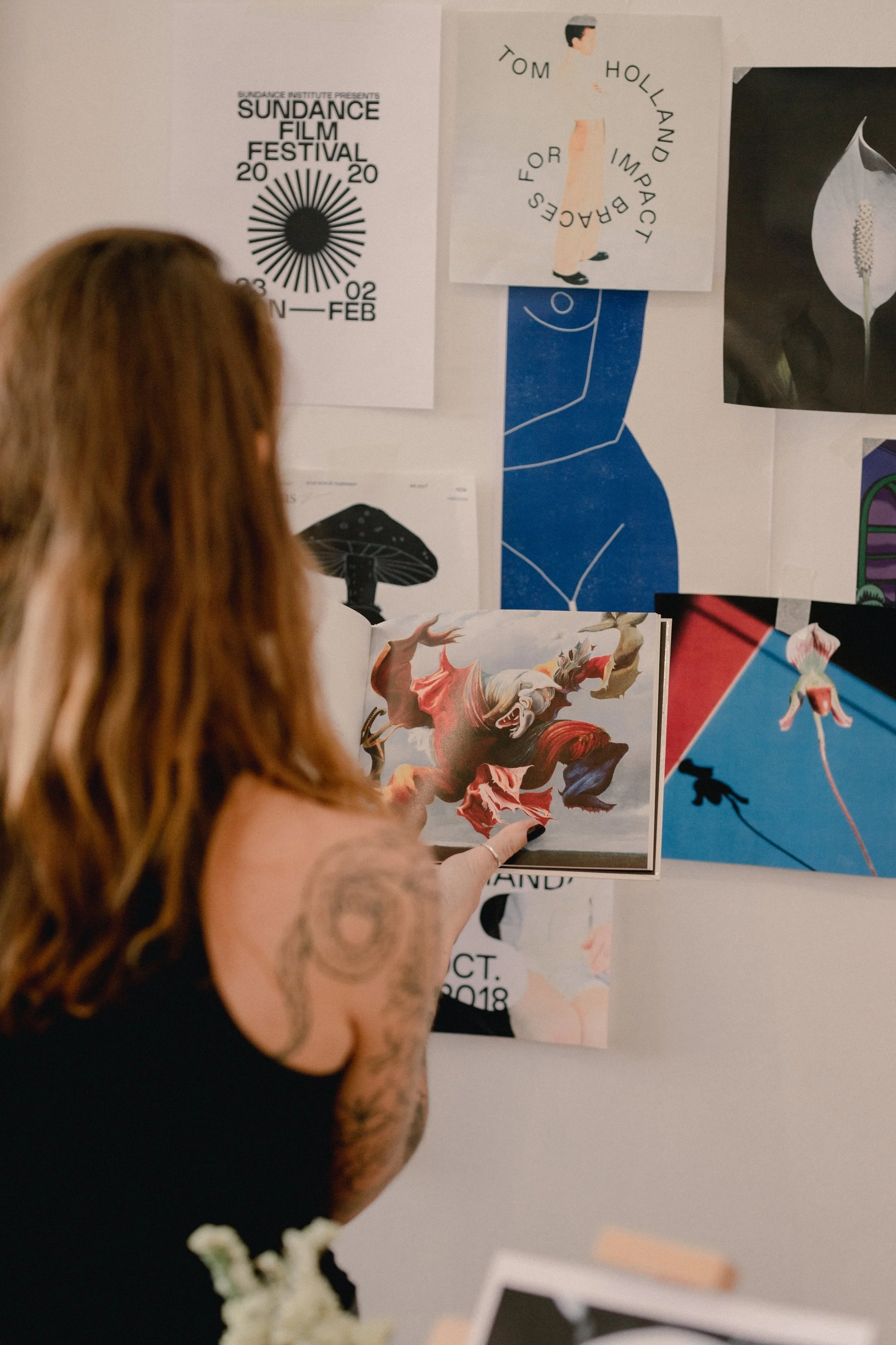 A woman with tattoos on her arm is pointing at a colorful picture of a lion on a wall decorated with various posters and artworks, including a film festival poster, a diagram of a face, a flower, and abstract art.