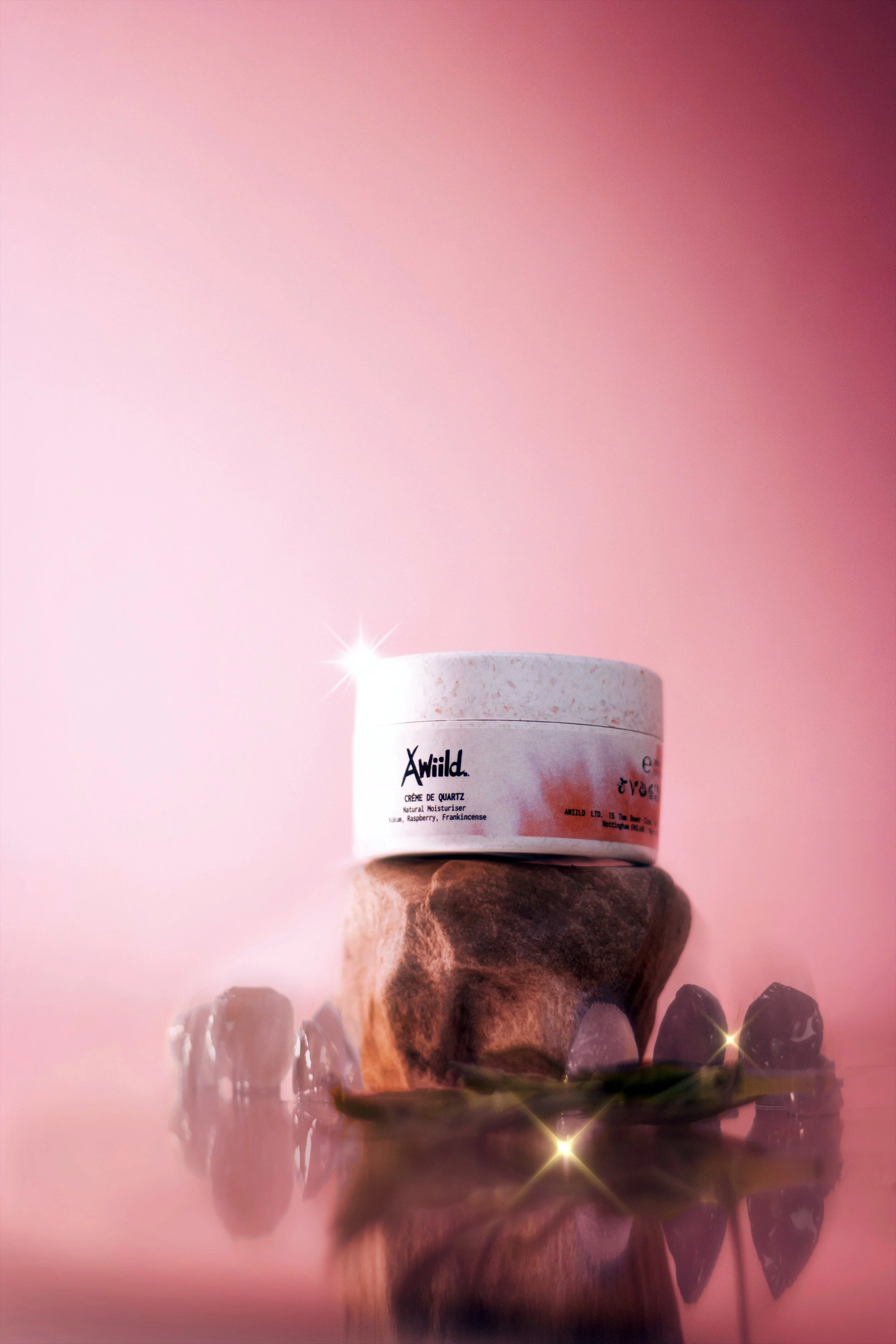 A skincare product container labeled 'Awild' positioned on a stack of rocks, with purple and white stones surrounding it, set against a pink background with a reflective surface.