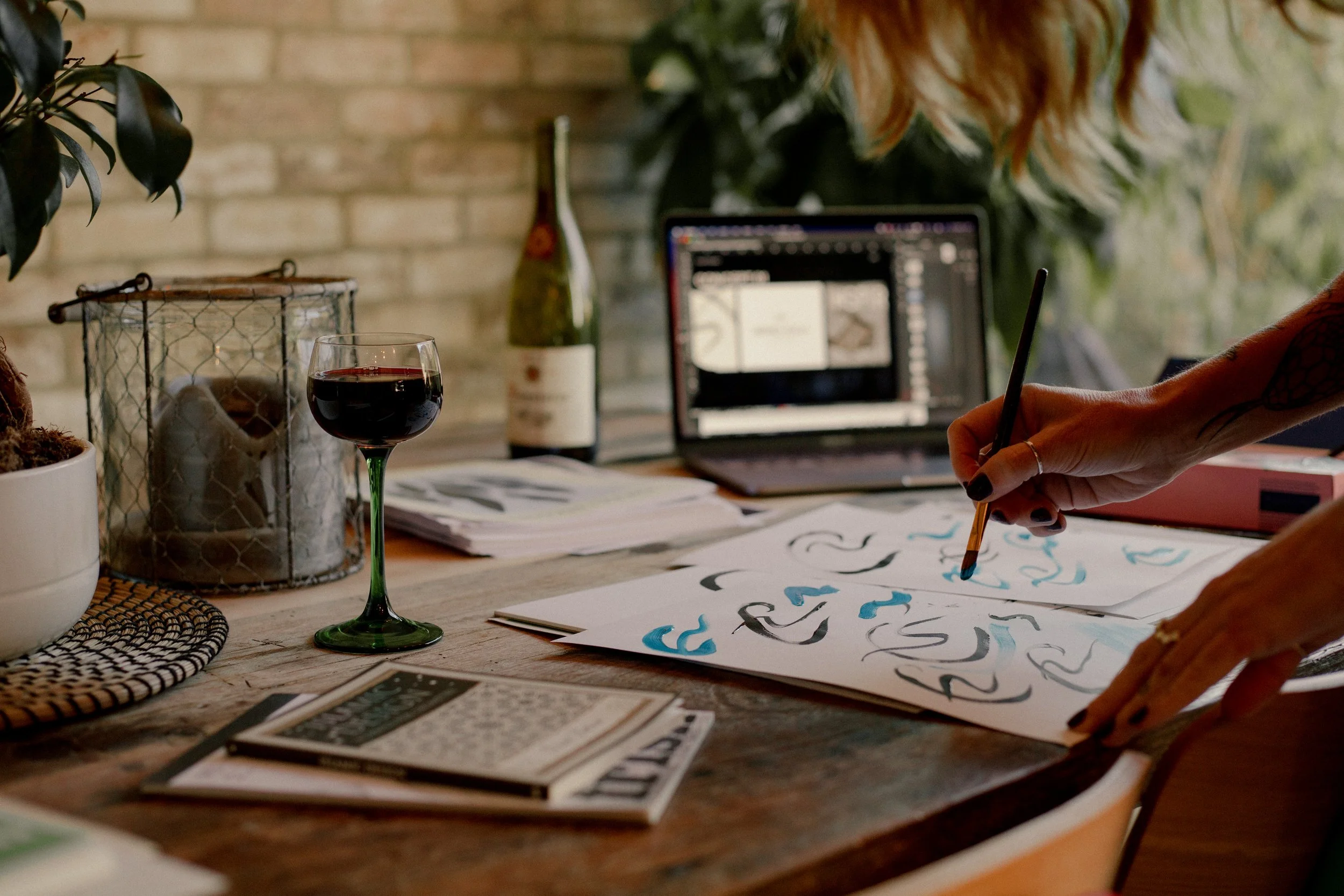 Person painting abstract patterns on paper at a cluttered wooden table with a glass of red wine, a bottle of wine, and a laptop in the background.