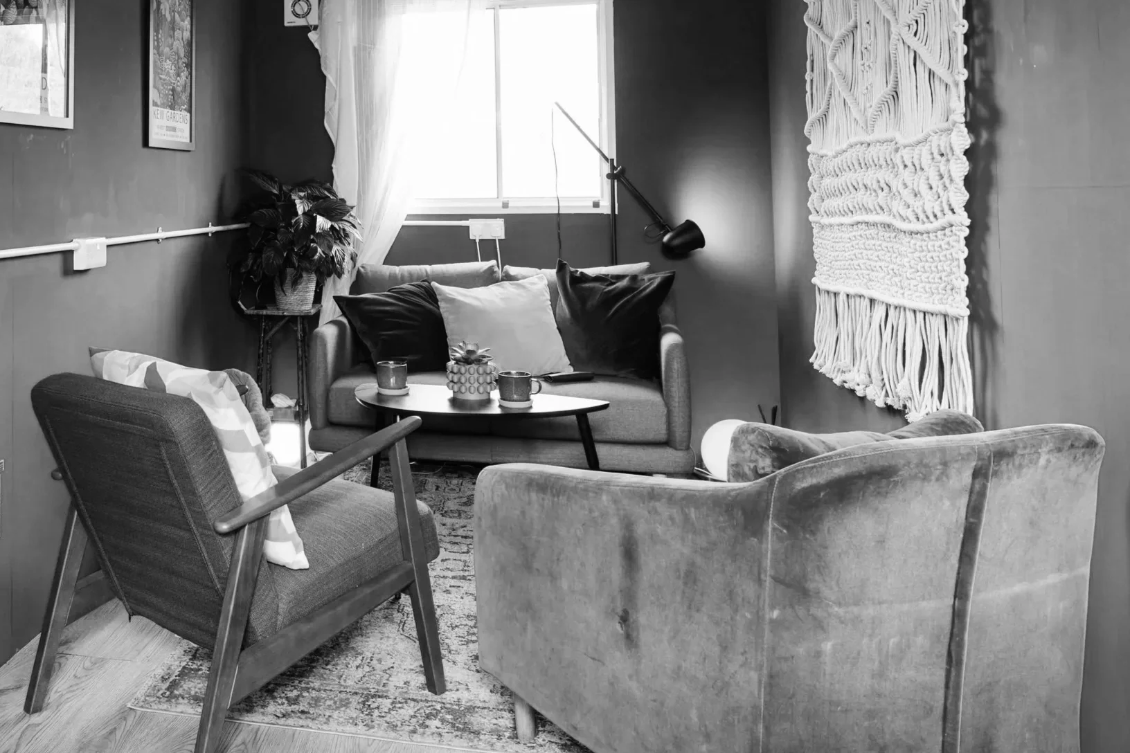 A cozy living room with a sofa, armchair, and a coffee table, decorated with pillows, candles, and a plant. There is a large window with white curtains, a wall hanging, and a floor lamp. The image is in black and white.