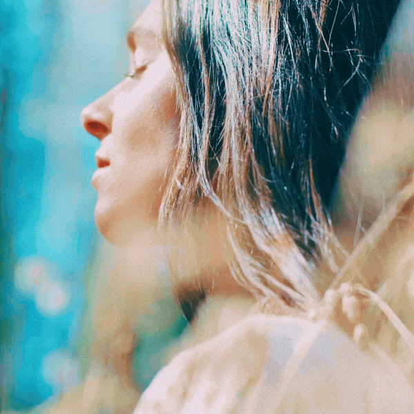 Close-up of a woman relaxing outdoors with her eyes closed, head tilted slightly, with sunlight and greenery in the background.