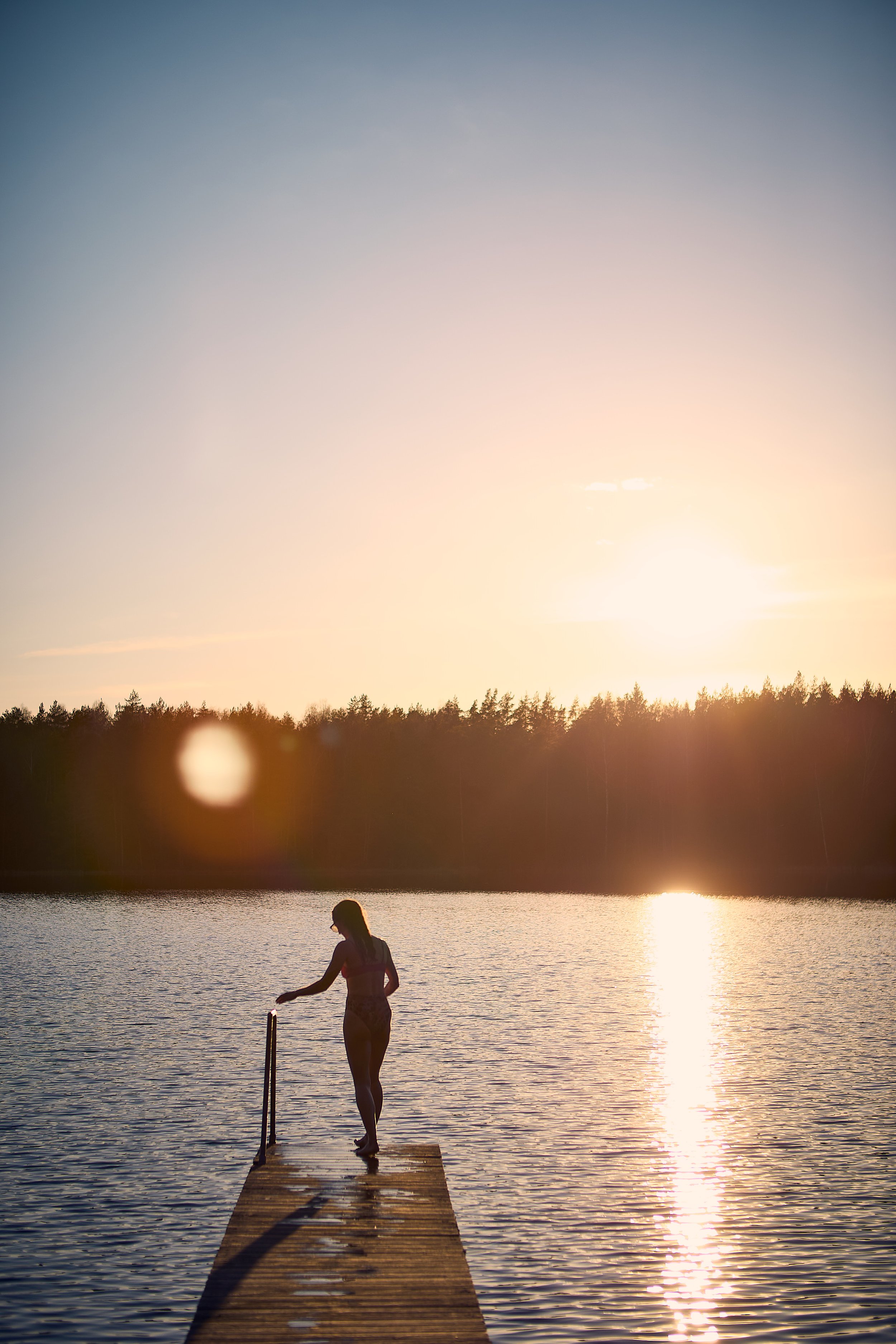 A woman stands on a wooden dock at sunset, reaching down to touch the water, with trees in the background and the sun reflecting off the lake.
