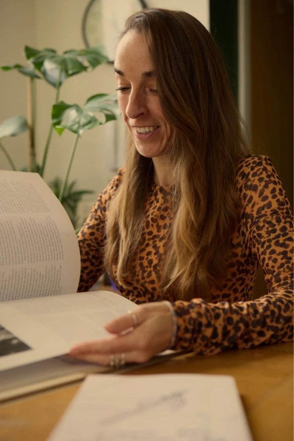 A woman with long brown hair wearing a leopard print shirt, sitting at a table, smiling while reading a book. There is a large green leafy plant in the background.
