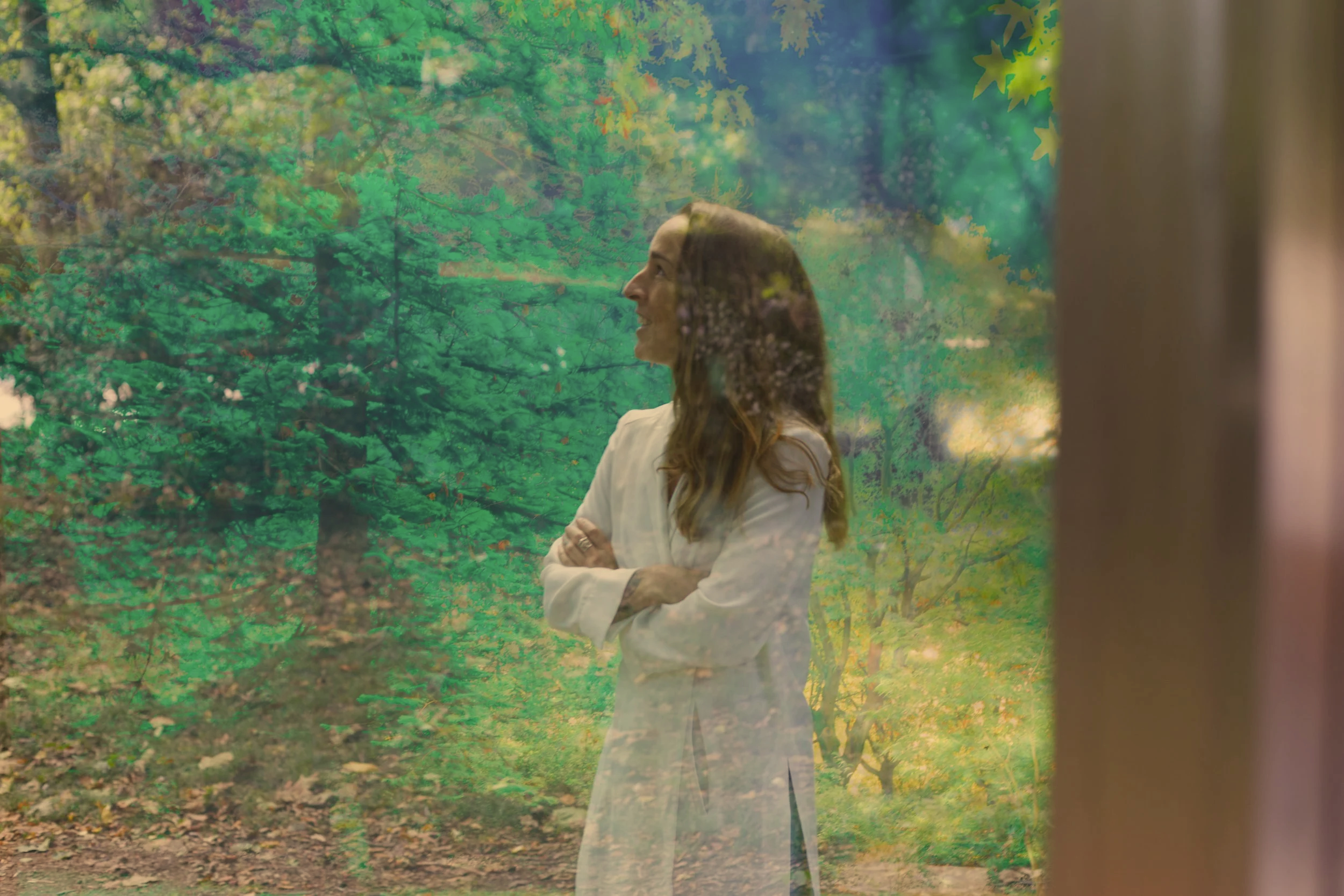A woman with long, wavy hair and a white blazer smiling with her arms crossed, looking to the right, standing outdoors near a wooded area with green leaves, behind a glass window.